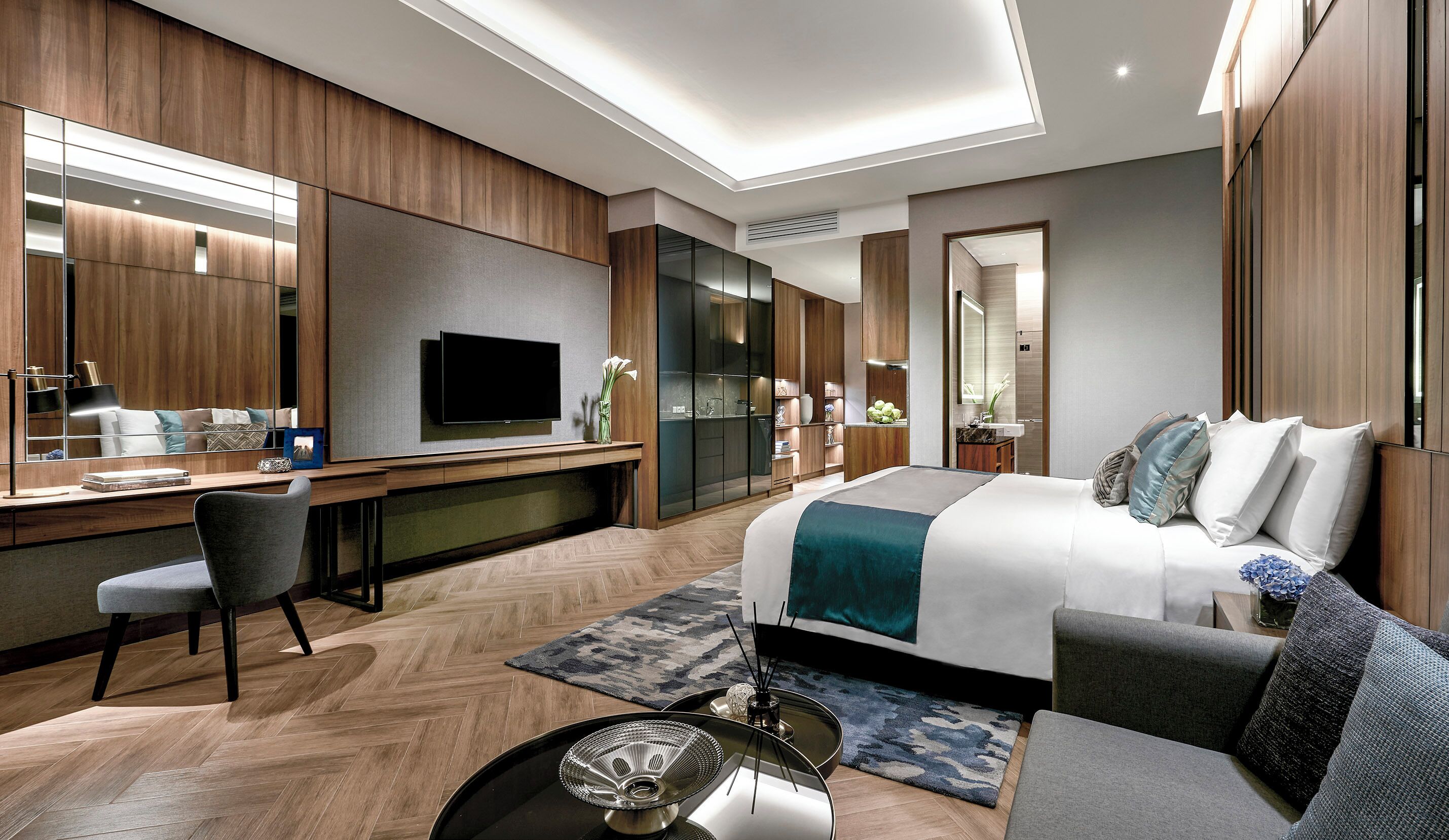 Una habitación de hotel moderna con una cama grande, un escritorio con televisor de pantalla plana, una zona de estar con sofá y sillones, y un espejo amplio.