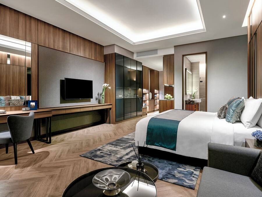 Una habitación de hotel moderna con una cama grande, un escritorio con televisor de pantalla plana, una zona de estar con sofá y sillones, y un espejo amplio.