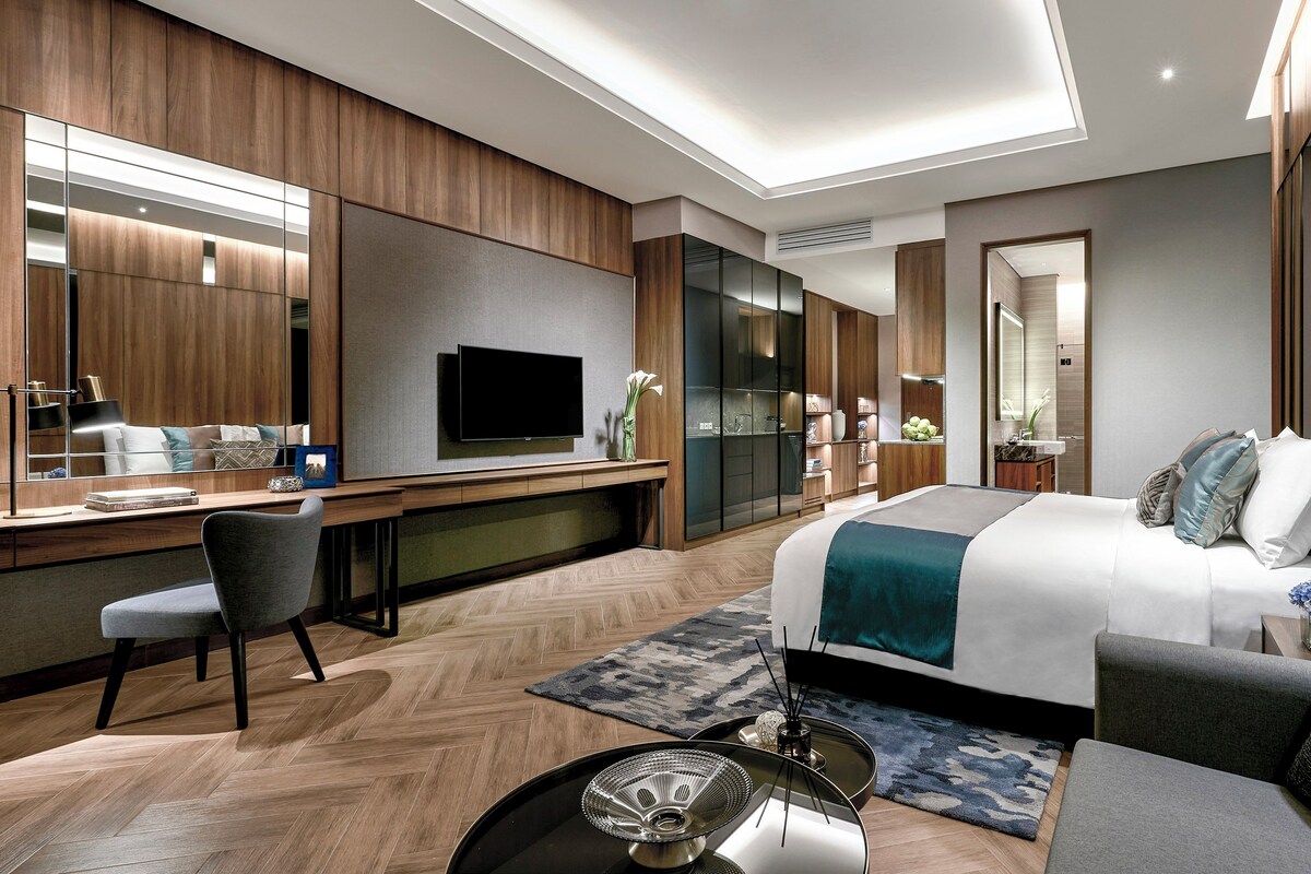 A modern hotel room with a large bed, a desk with a flat-screen TV, a seating area with a sofa and armchairs, and a large mirror.