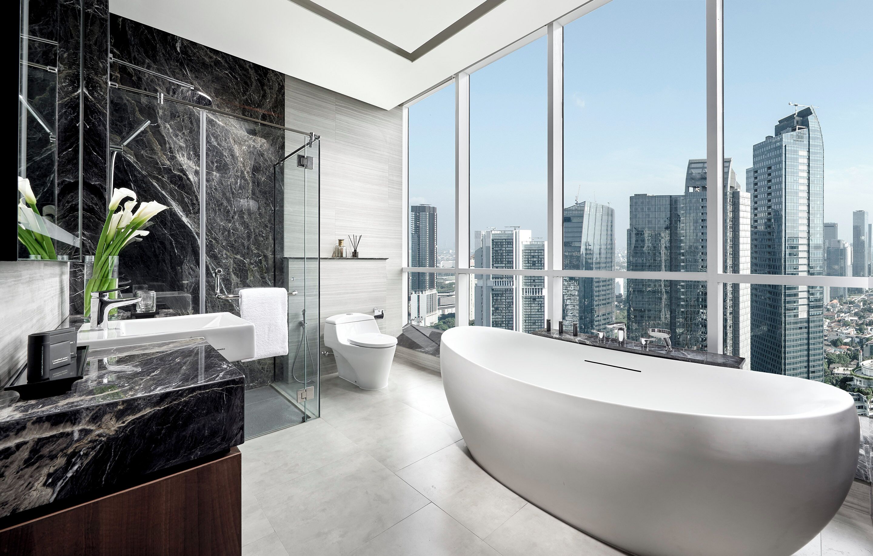 Un baño moderno con una bañera grande, una ducha con mampara de vidrio y un inodoro. Cuenta con una encimera de mármol y un ventanal con vistas a la ciudad.