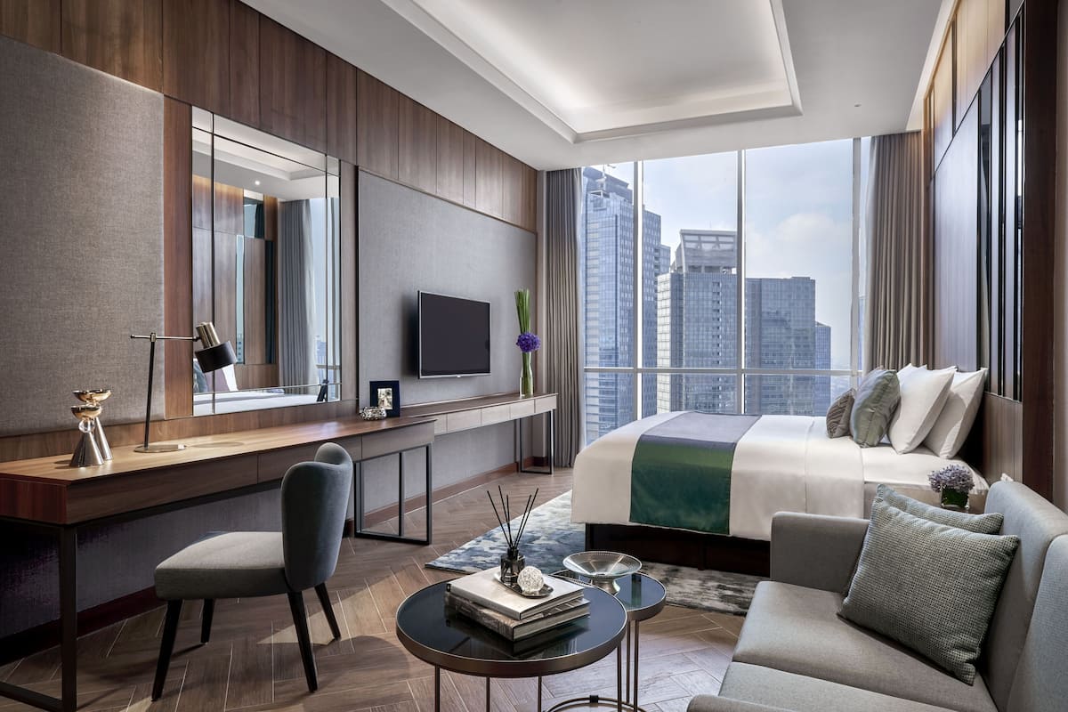 A modern hotel room with a large bed, a desk with a flat-screen TV, a comfortable sofa, and a view of the city skyline.
