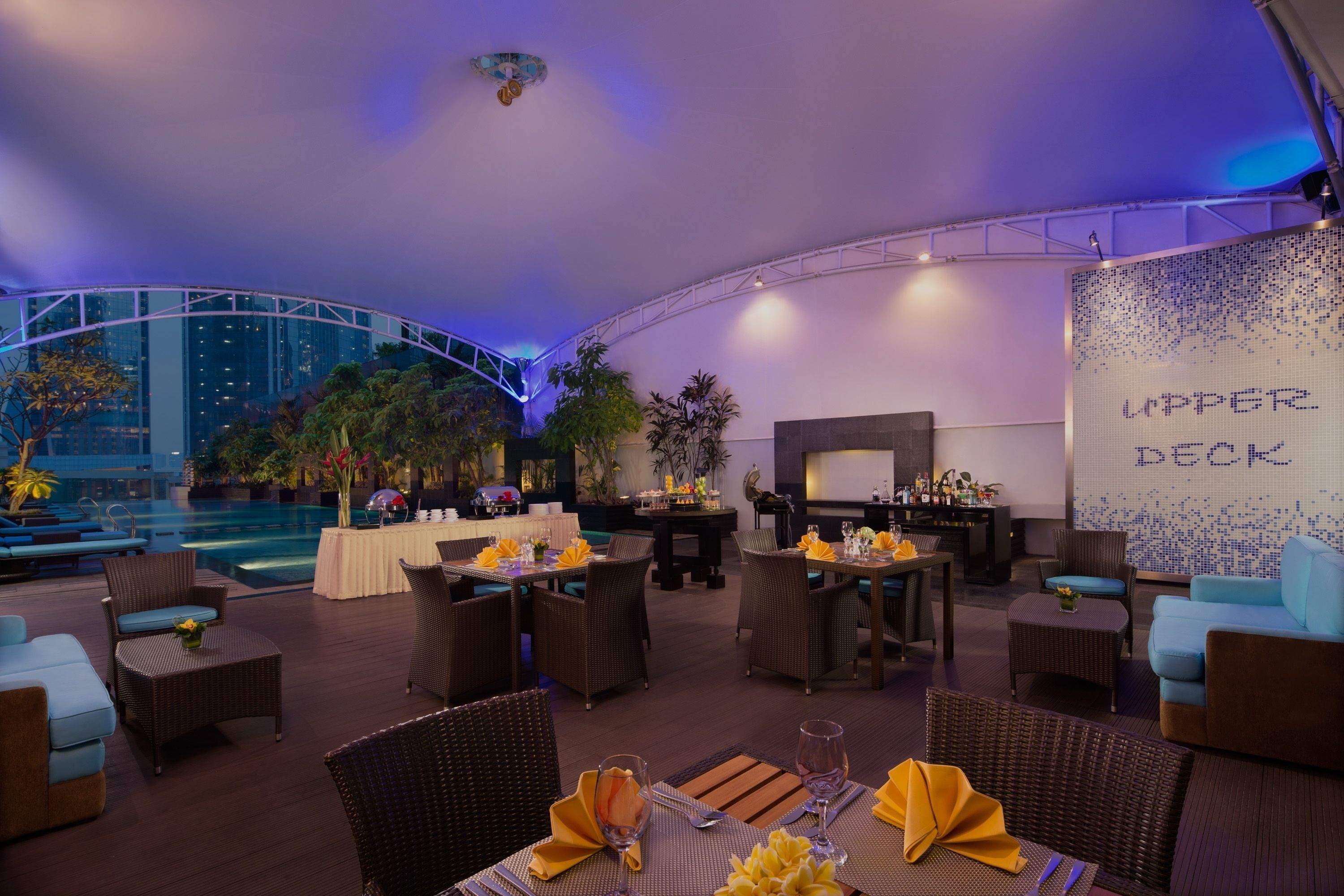 A rooftop lounge with dining tables, wicker furniture, and a view of the city skyline.