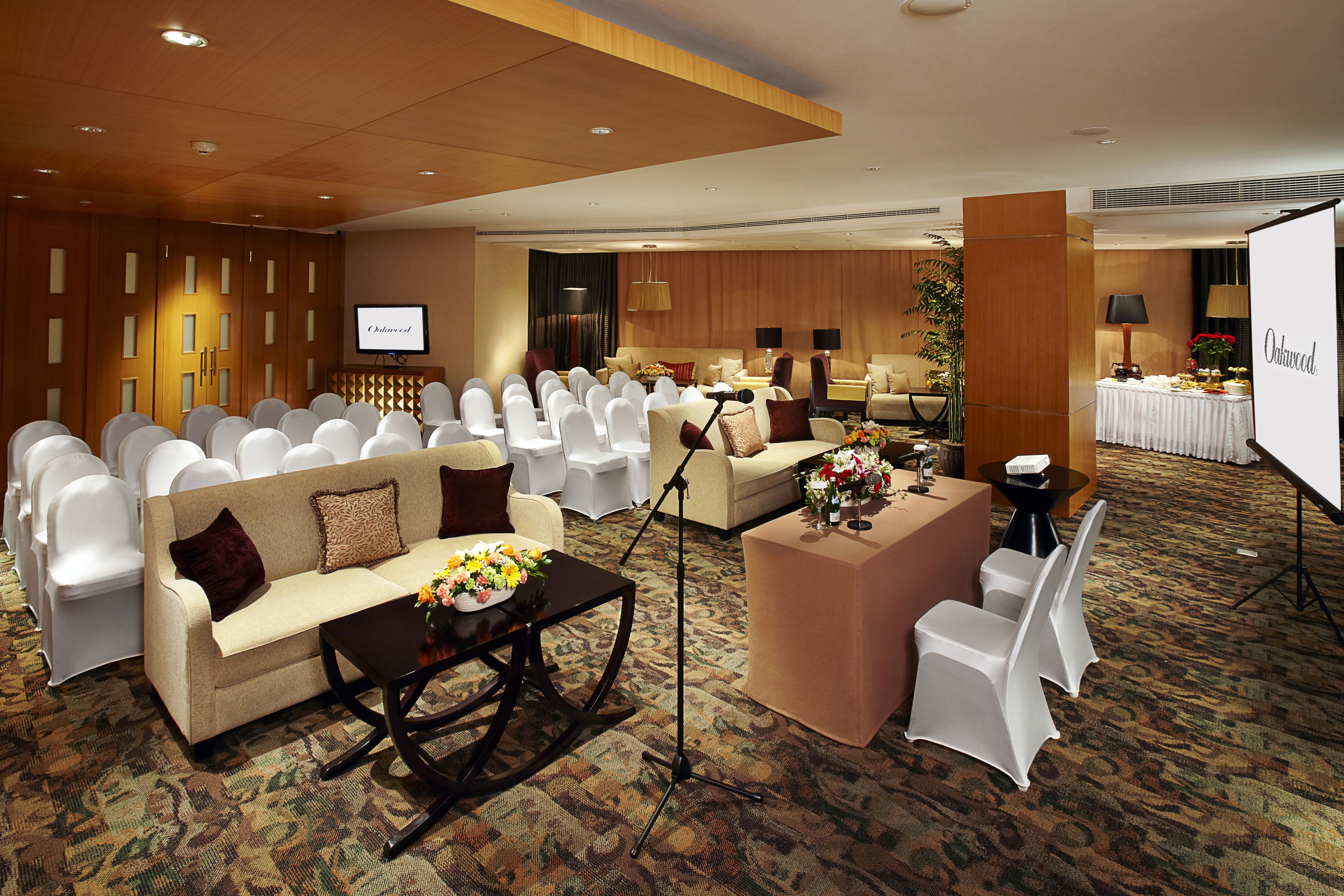A conference room with rows of white chairs, a sofa, a coffee table with flowers, and a projection screen.