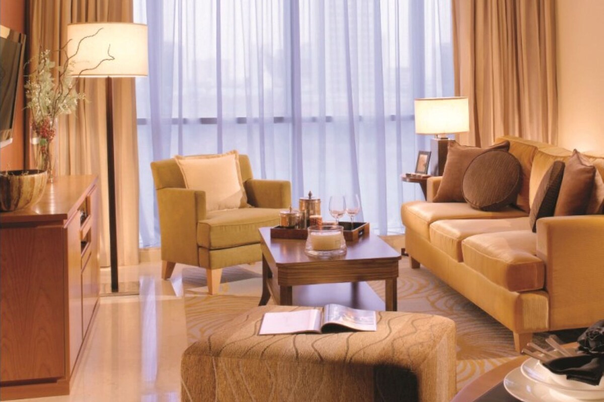 A hotel room with a sofa, armchair, coffee table, and a large window with curtains.
