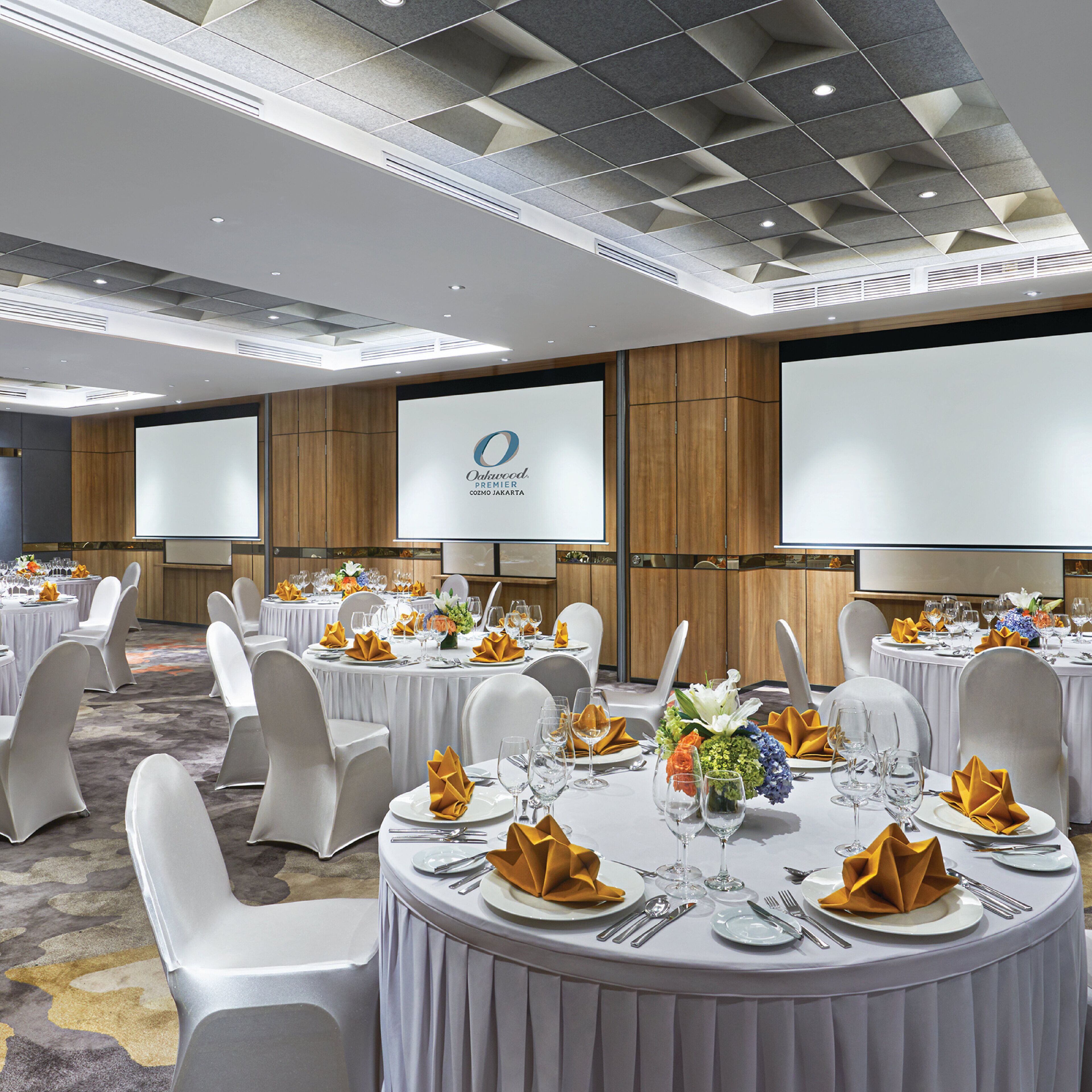 A conference room with multiple tables set for a formal event, equipped with chairs, tables, and a large screen displaying a logo.