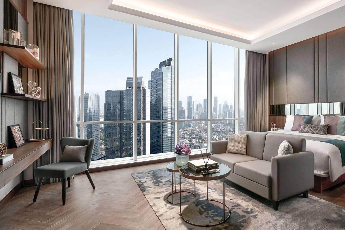 A modern hotel room with a large window, a sofa, a bed, a TV, and a small table with a vase of flowers.