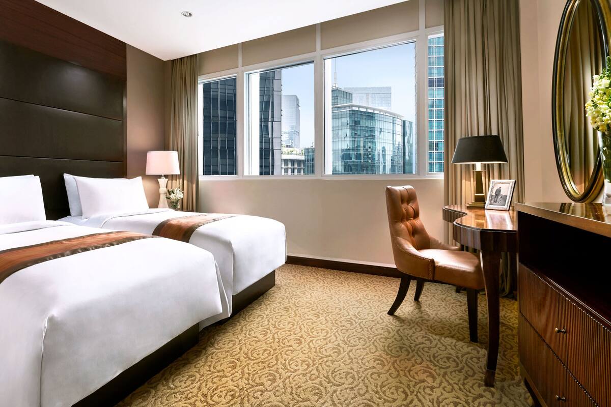 A hotel room with a large bed, a desk, a chair, and a view of the city through large windows.