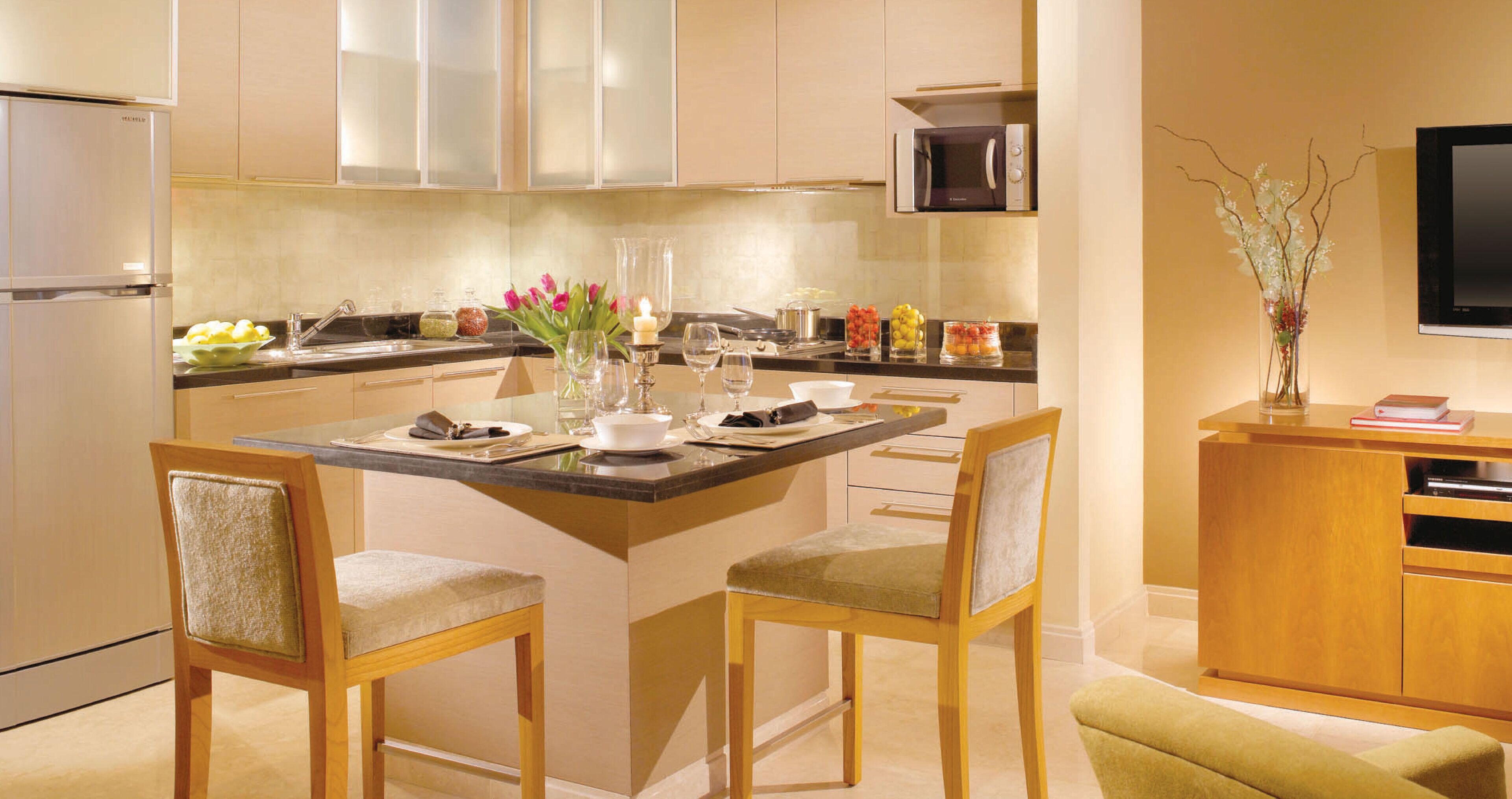 A modern kitchen with a central island, wooden chairs, a microwave, and a flat-screen TV.