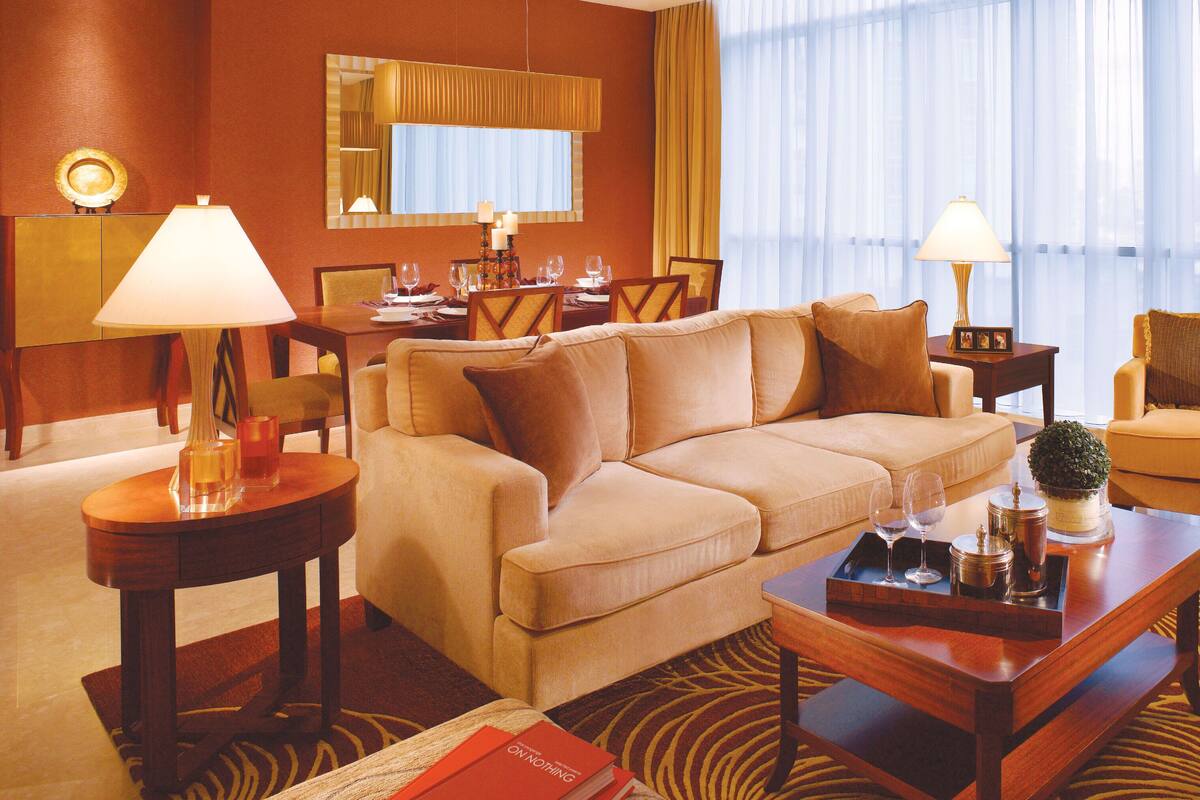 A well-lit hotel room with a beige sofa, a dining table set for four, a small side table, and a large window with sheer curtains.