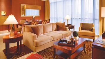 A well-lit hotel room with a beige sofa, a dining table set for four, a small side table, and a large window with sheer curtains.