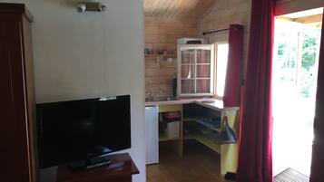 Een compacte kamer met een televisie op een houten standaard, een klein raam met uitzicht op groen en een kitchenette met een magnetron en wastafel.