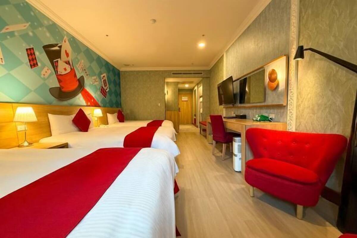 A hotel room with three beds, a desk, a TV, and a red armchair.
