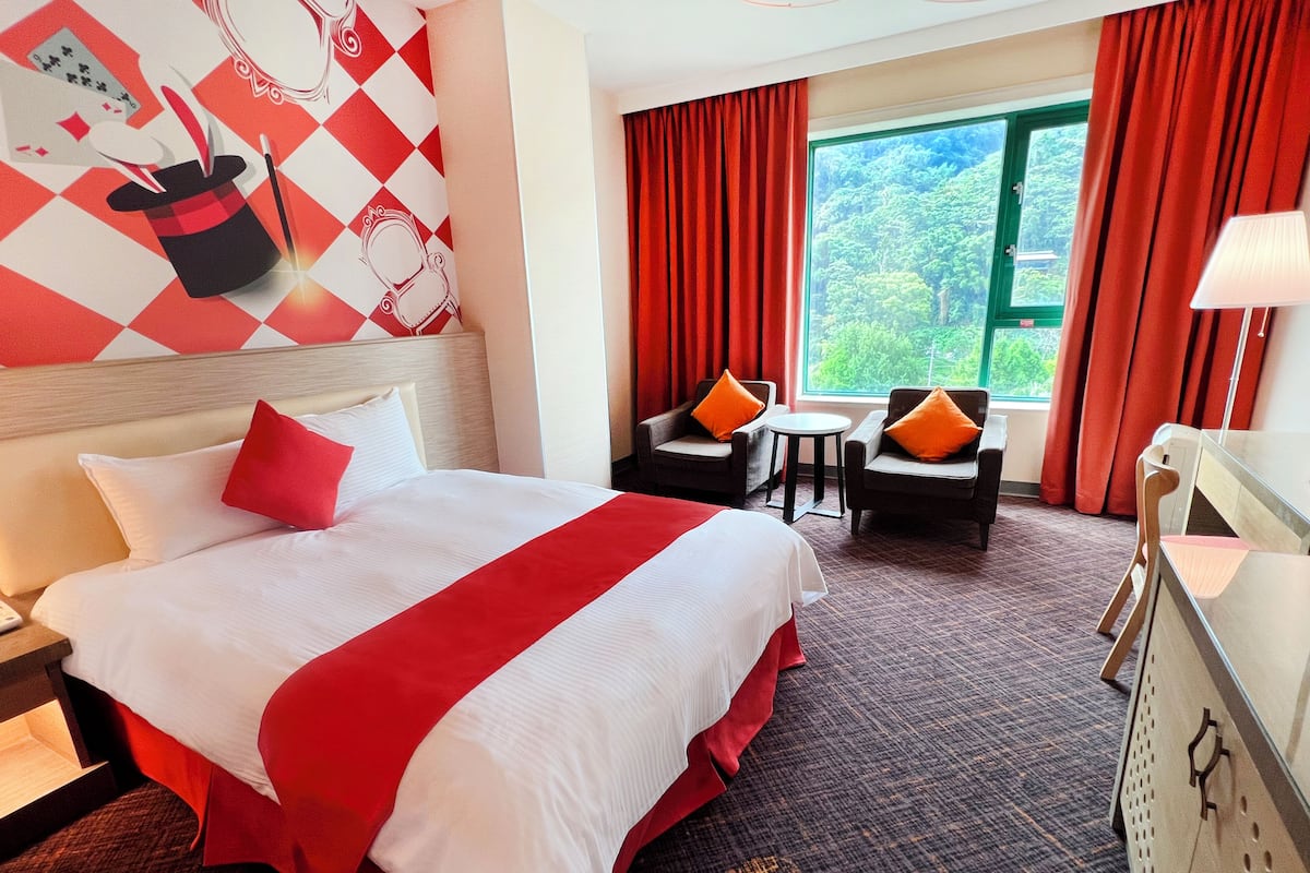 A hotel room with a large bed, two armchairs, a small table, and a view of greenery through the window.
