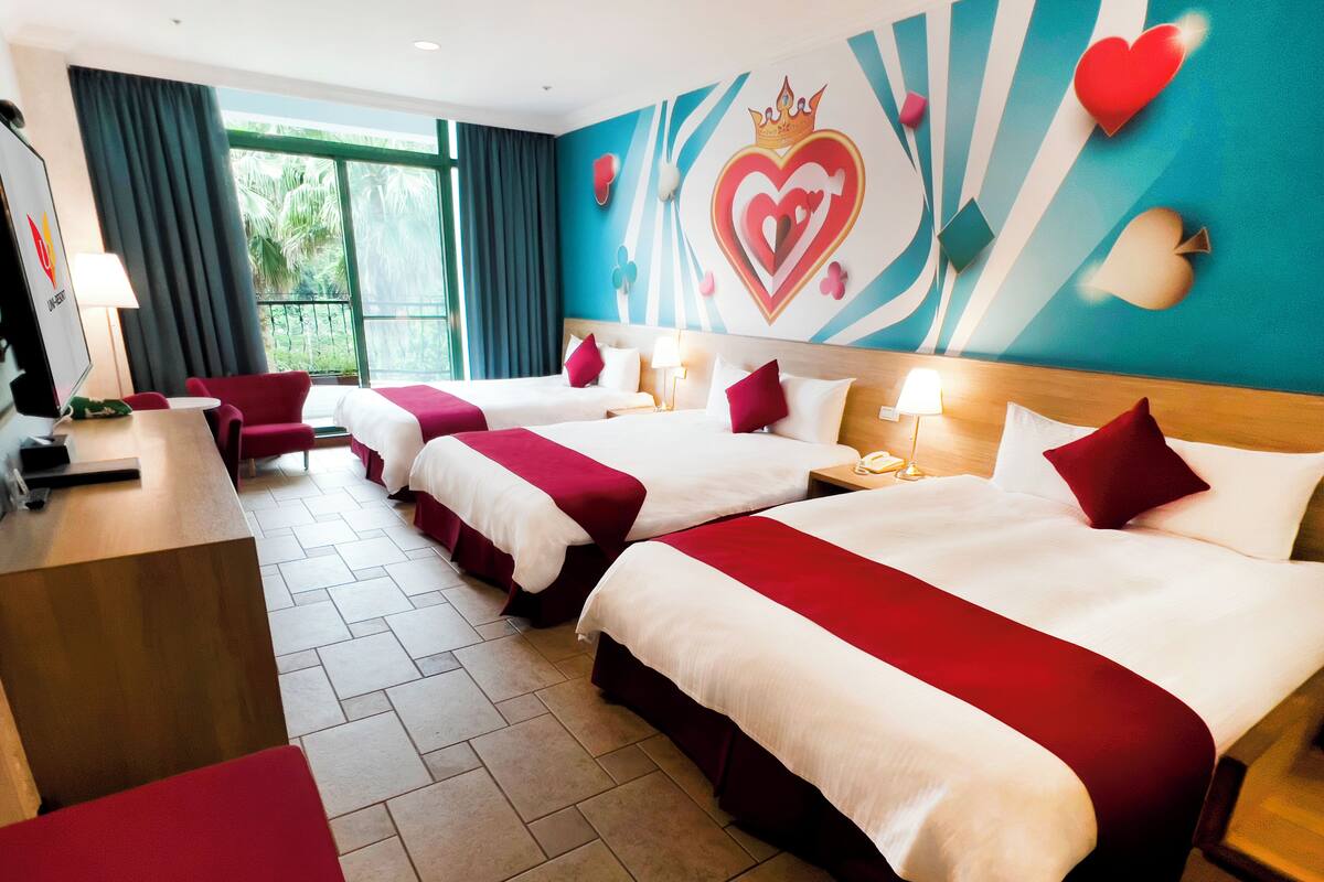 A hotel room with four beds, a desk, and a view of palm trees through the window.