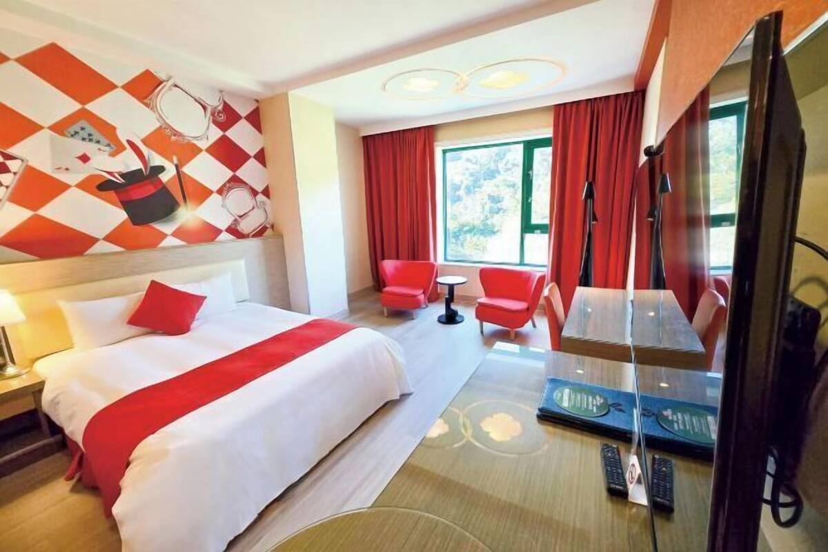 A hotel room with a bed, red chairs, a small table, a TV, and a window with curtains.