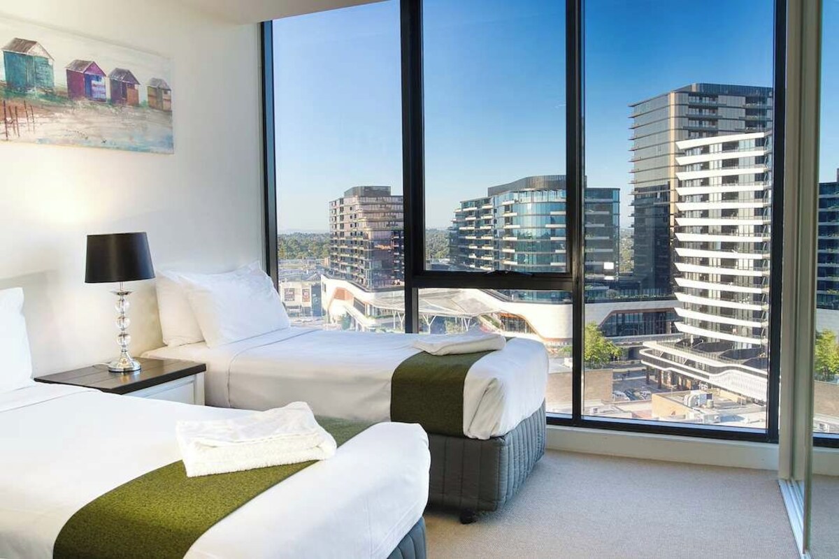 A hotel room with a large window, a bed, a bedside table with a lamp, and a painting on the wall.