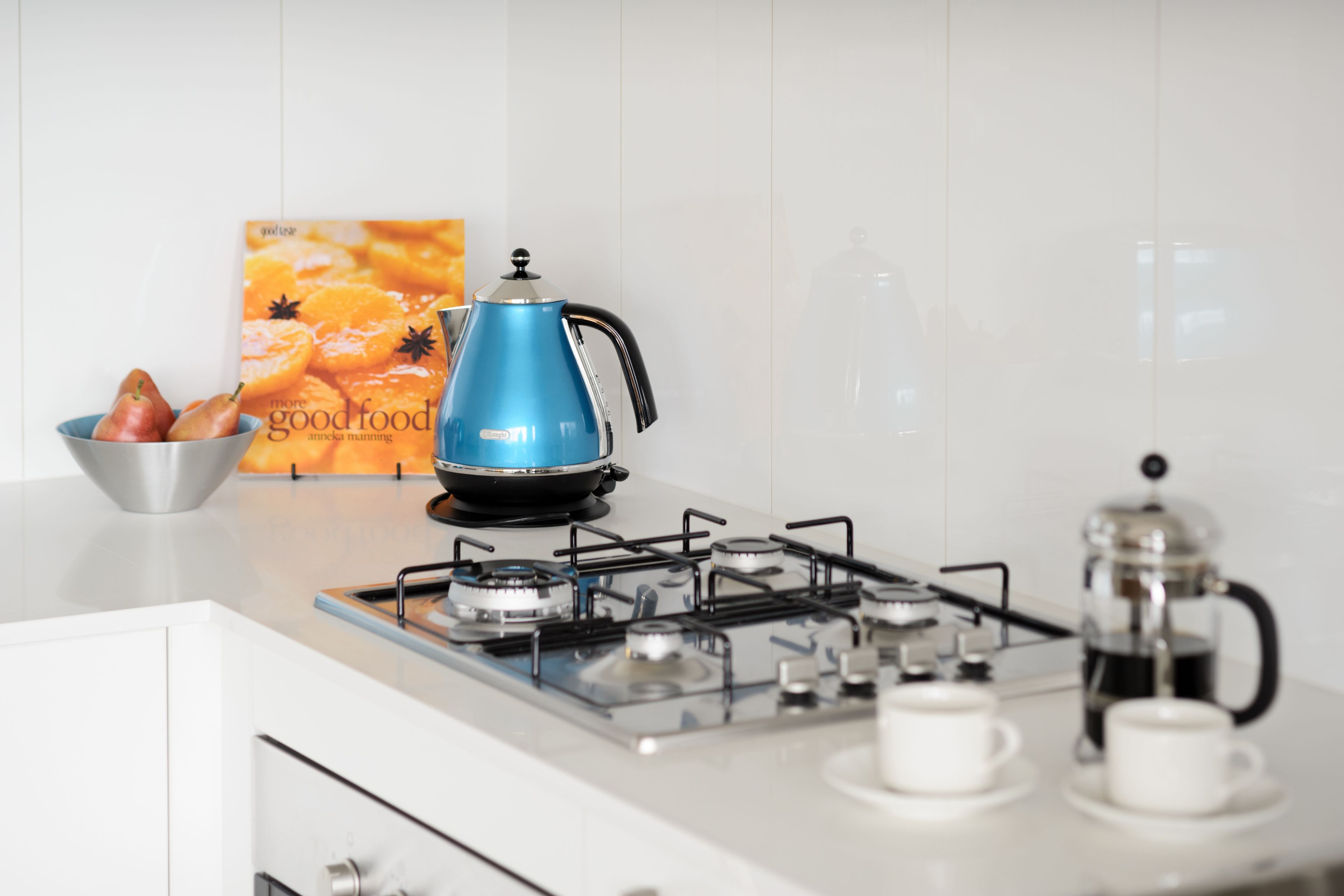 Eine Küche mit einem Gasherd, einem blauen elektrischen Wasserkocher, einer Kaffeemaschine und einer Schüssel mit Obst.