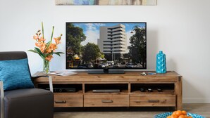 A living room with a flat-screen TV on a wooden TV stand, a sofa with a blue cushion, a vase with flowers, and a bowl of oranges on a coffee table. - Park Avenue - IKON Glen Waverley (Glen Waverley)