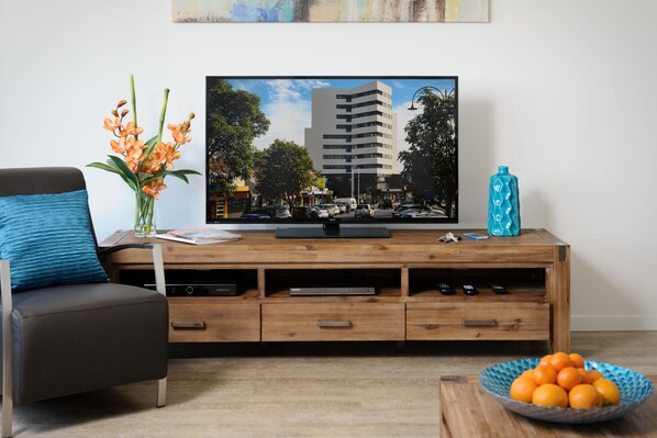 A living room with a flat-screen TV on a wooden TV stand, a sofa with a blue cushion, a vase with flowers, and a bowl of oranges on a coffee table. - Park Avenue - IKON Glen Waverley (Glen Waverley)