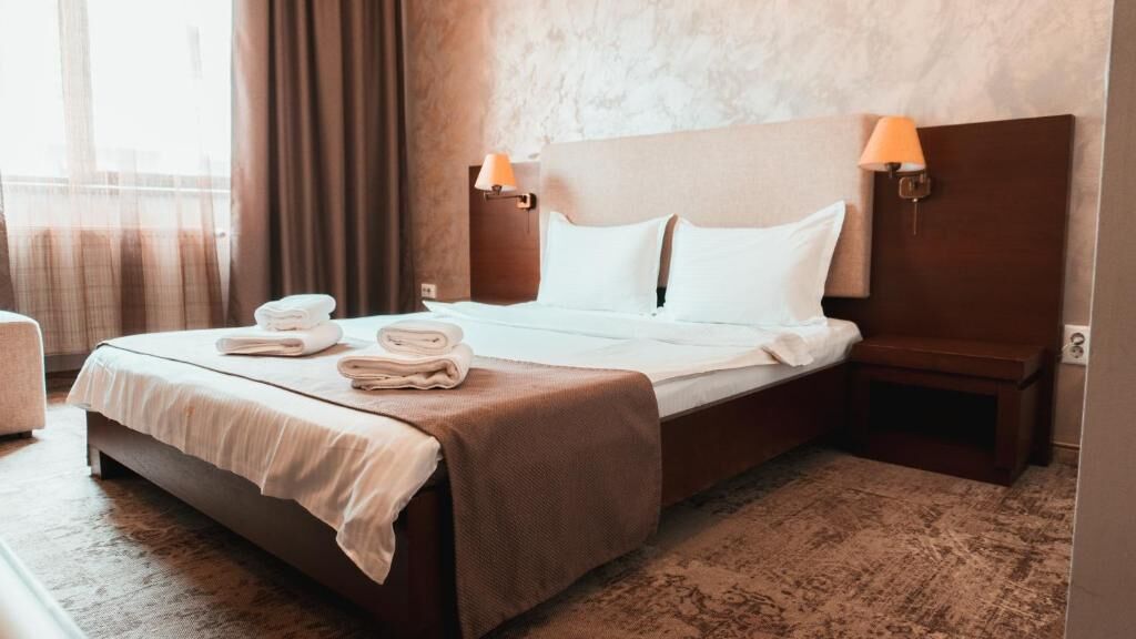 A hotel room with a bed, two bedside lamps, a bedside table, and a window with curtains.