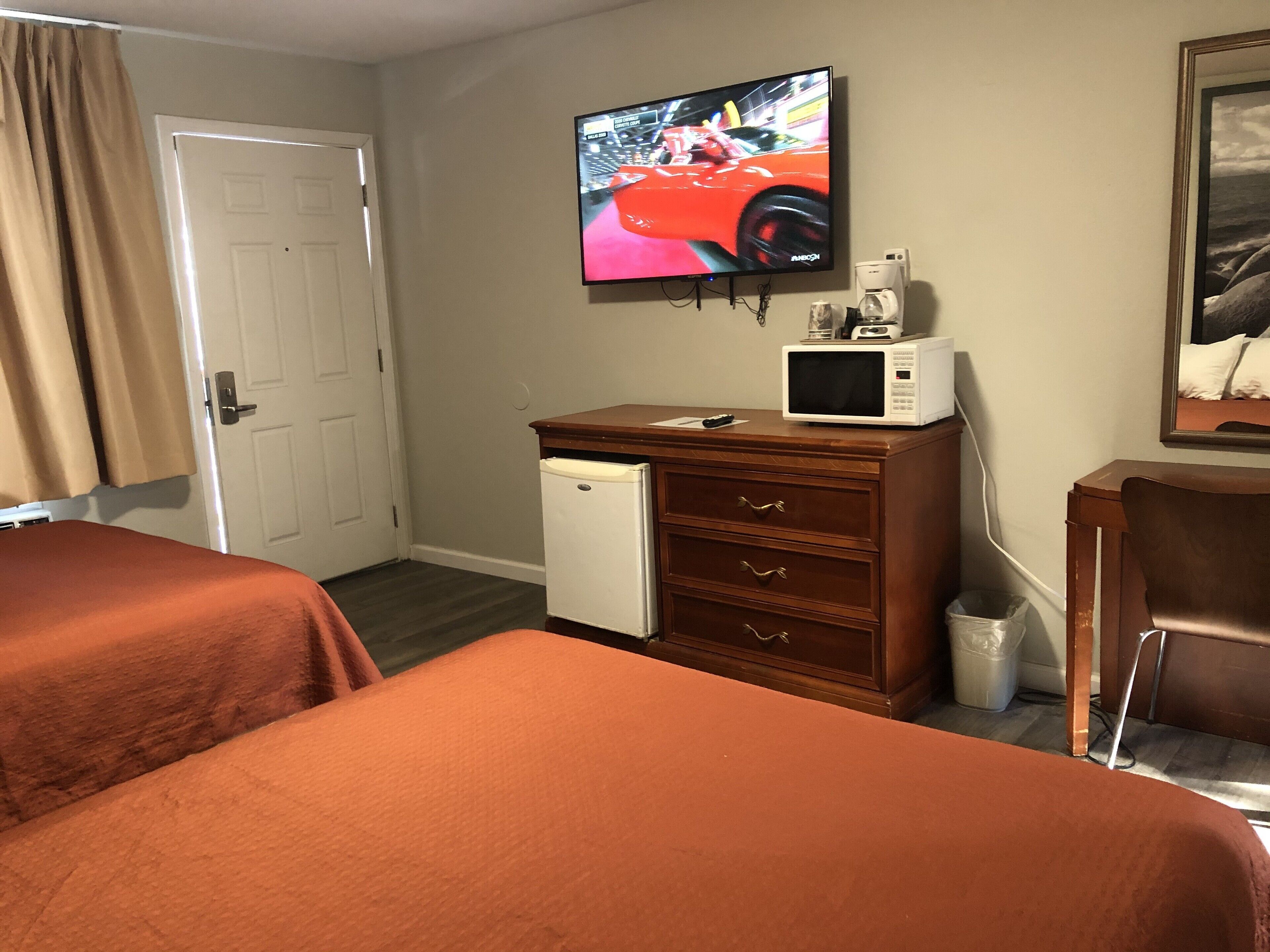 Habitación de hotel con dos camas, un televisor montado en la pared, un refrigerador pequeño y un cuadro decorativo.