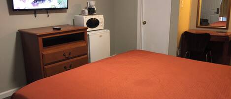 A bedroom with a bed, a dresser, a coffee maker, a refrigerator, a television, and a mirror.