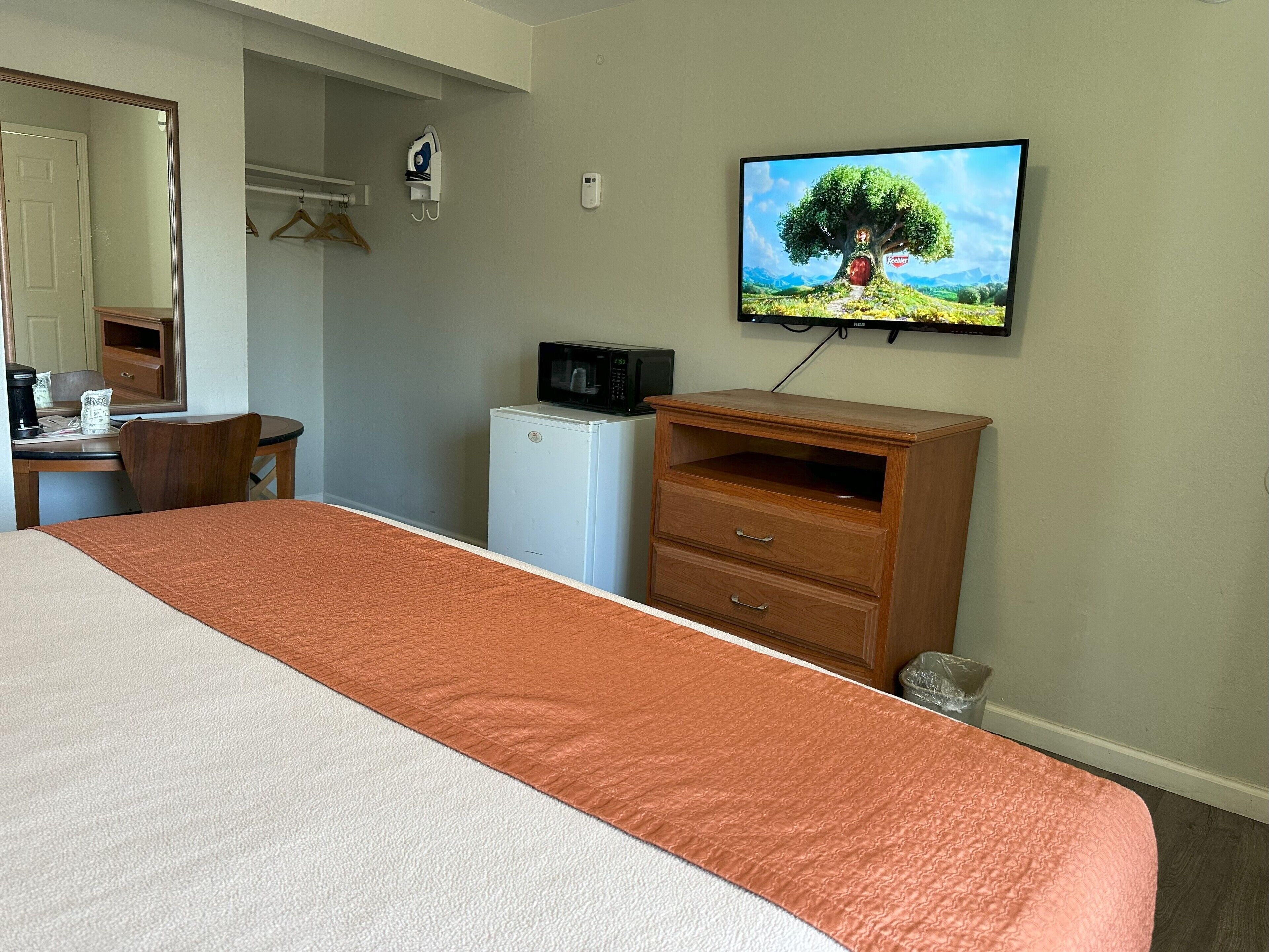 A hotel room with a bed, a desk with a computer, a microwave, a TV, and a small refrigerator.