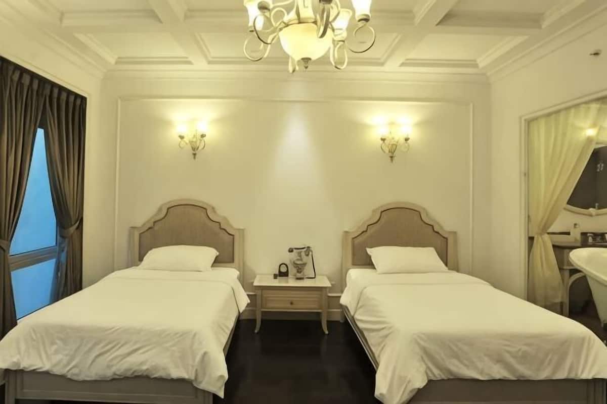 A hotel room with two beds, a nightstand, a chandelier, wall sconces, a window with curtains, and a bathroom mirror.