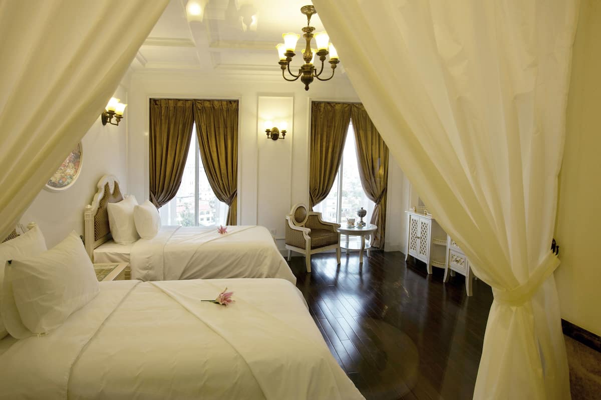 A hotel room with two beds, a chandelier, a chair, and a small table with a lamp.