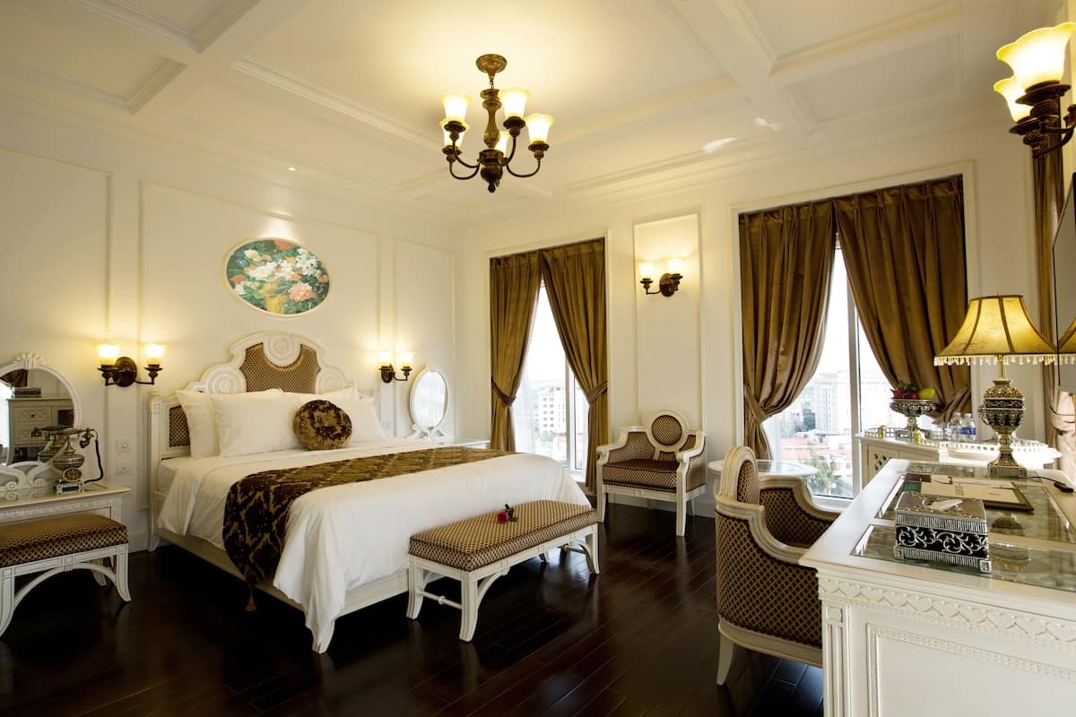 A spacious hotel room with a large bed, a sitting area with a chair and bench, a desk with a lamp, and a view of the city through the windows.