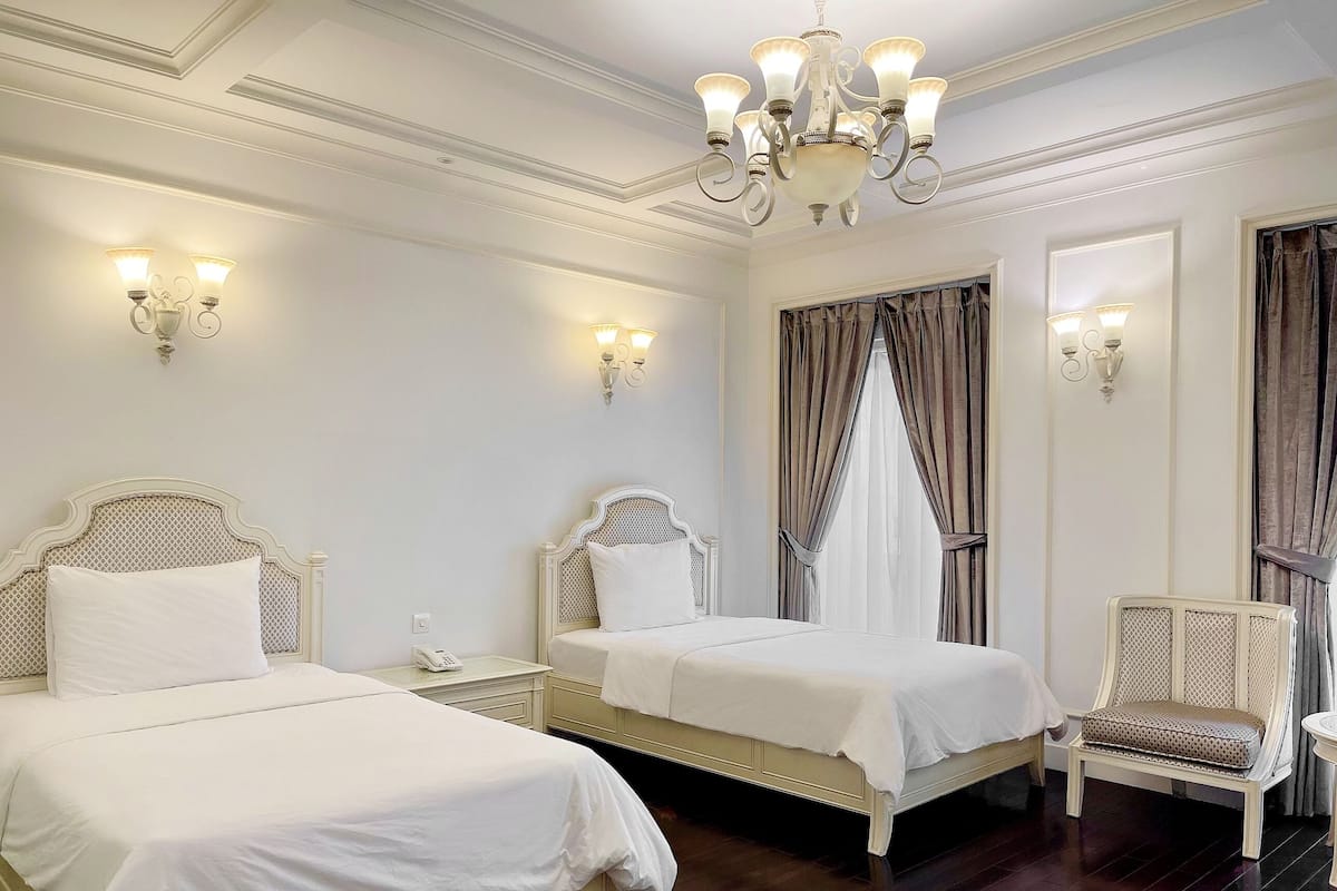 A hotel room with two beds, a chandelier, wall sconces, and a chair.