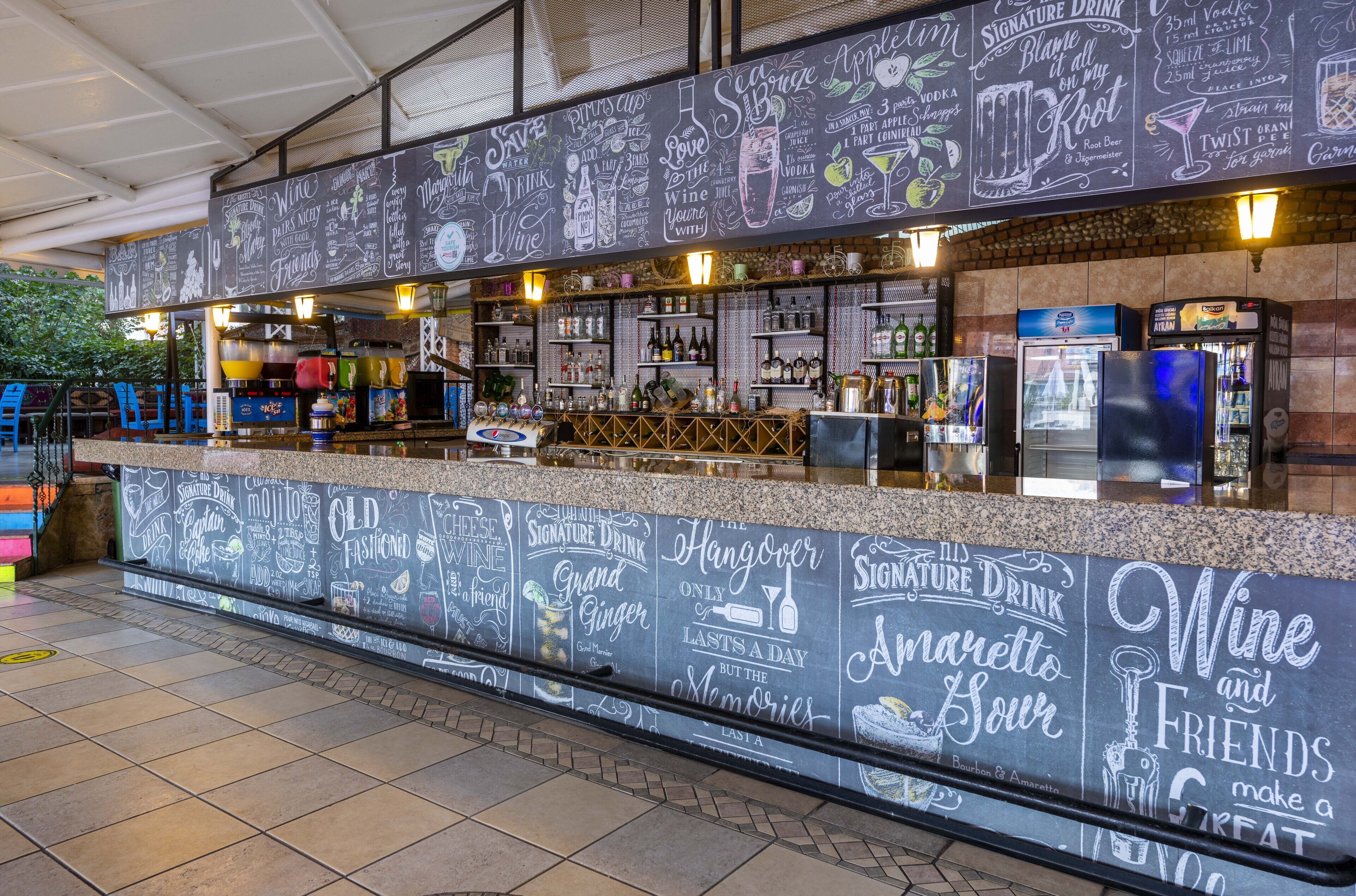 Un espace bar avec un mur recouvert d’un tableau noir où sont affichées différentes options de boissons, ainsi qu’un comptoir où sont présentées diverses bouteilles et verres.