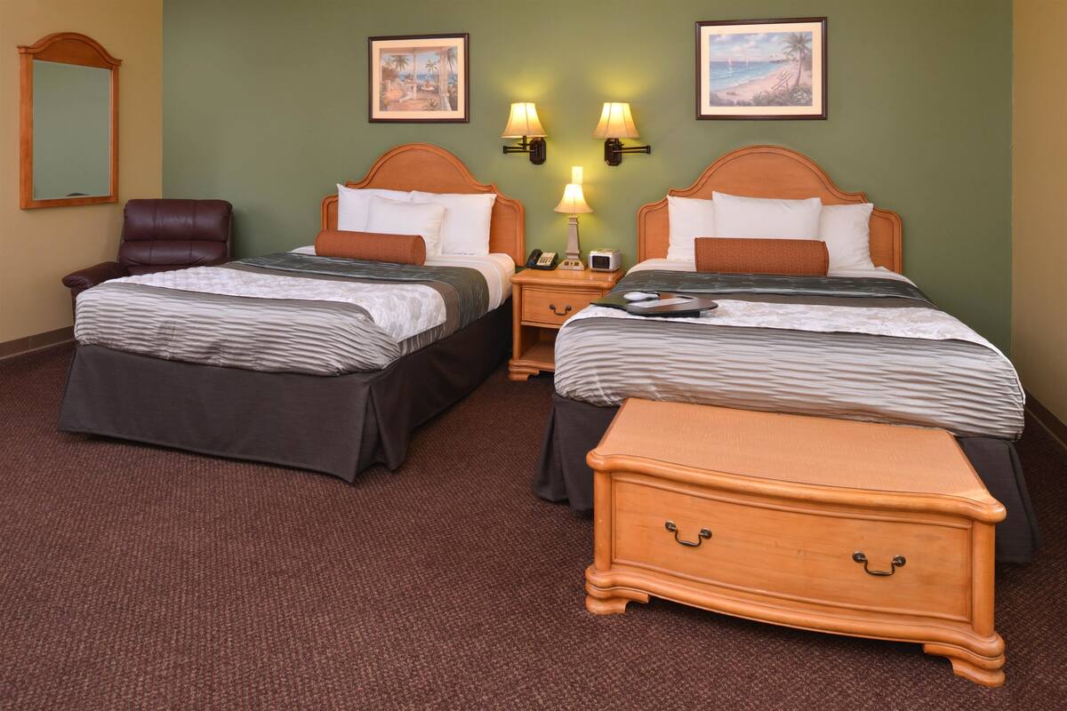 Une chambre d’hôtel avec deux lits, une table de chevet avec une lampe, une commode en bois, une chaise et deux lampes fixées au mur.