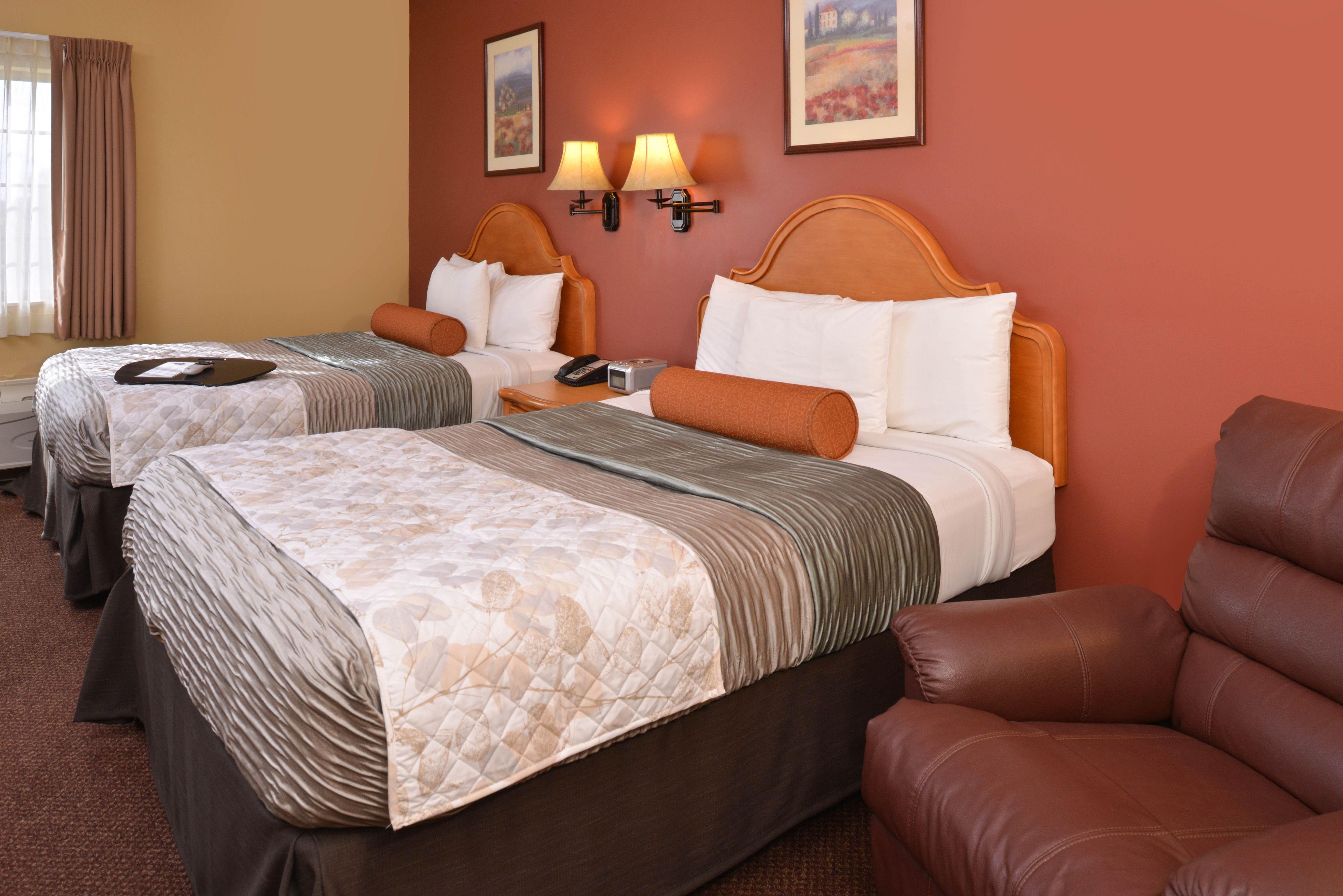 Quarto de hotel com duas camas, uma poltrona de couro marrom e duas luminárias fixadas na parede.
