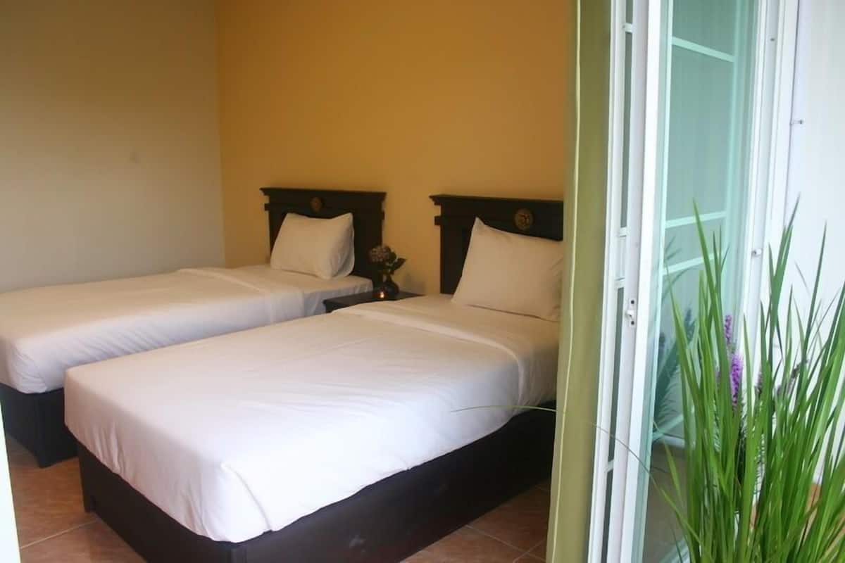 A hotel room with two single beds, a small table with a vase, and a sliding glass door leading to a balcony with plants.