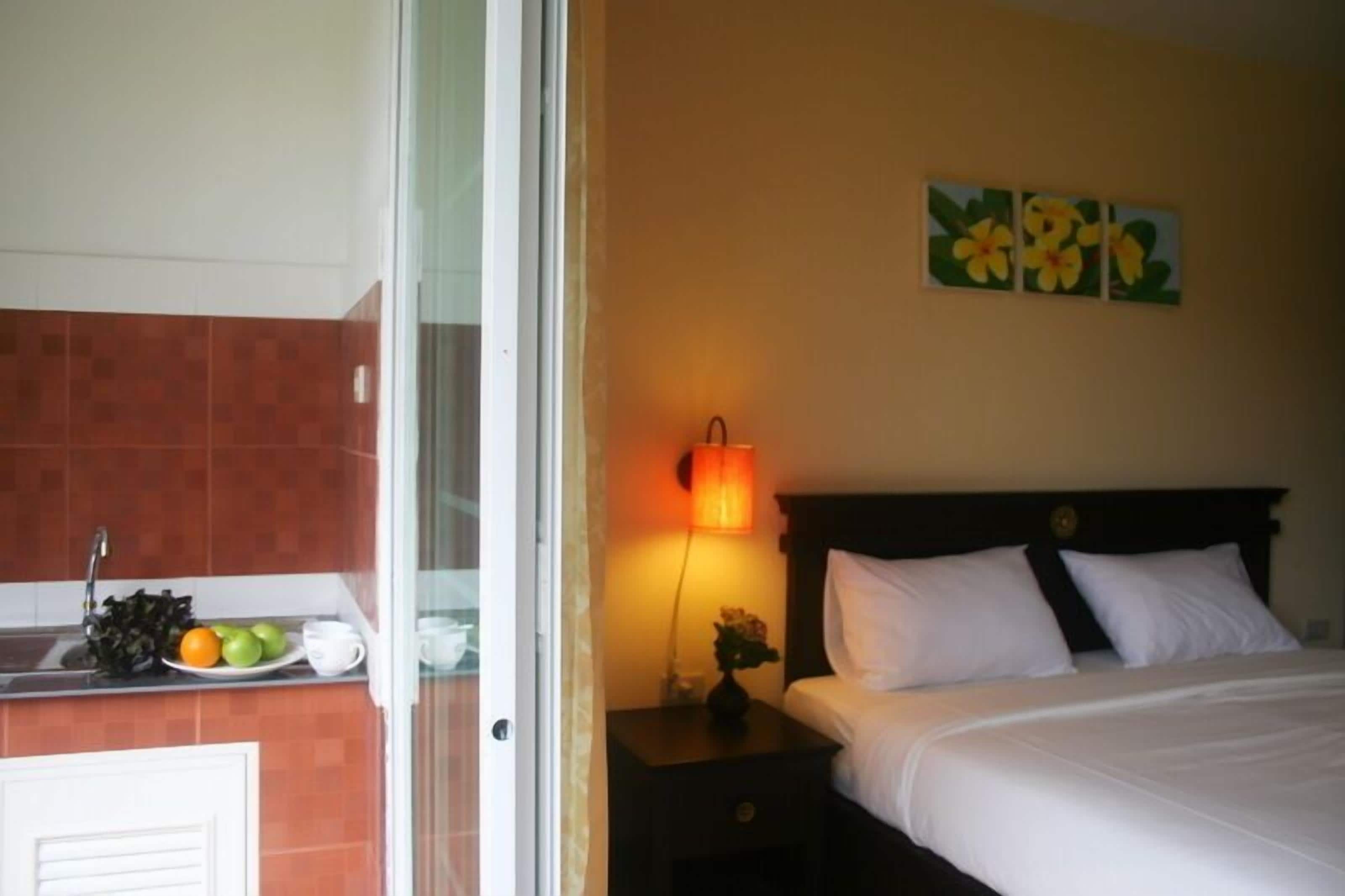 A hotel room with a bed, a nightstand with a lamp, a wall with floral artwork, a kitchen area with a sink and fruit bowl.