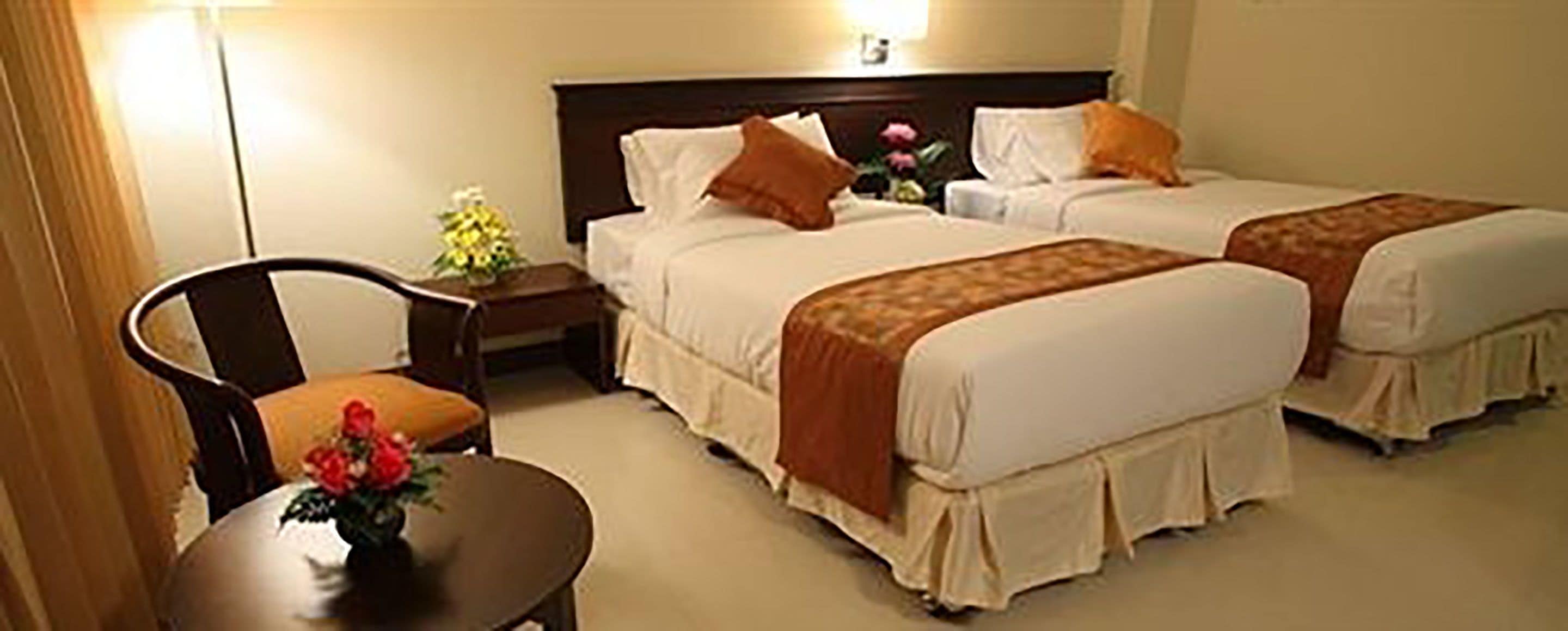 A hotel room with two beds, a chair, a small table, and a vase with flowers.