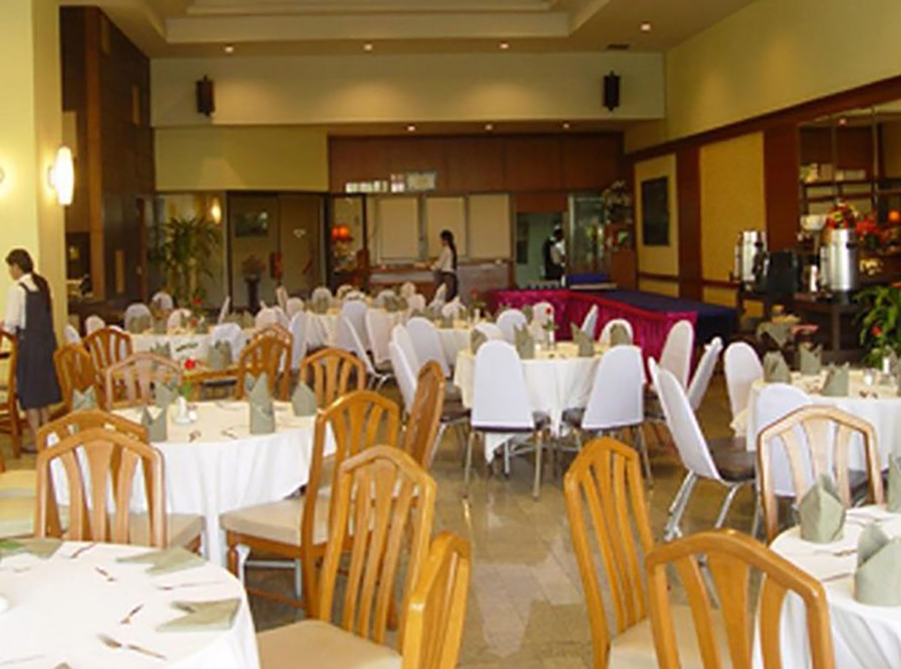 A banquet hall with round tables and wooden chairs, a stage, and a buffet area.