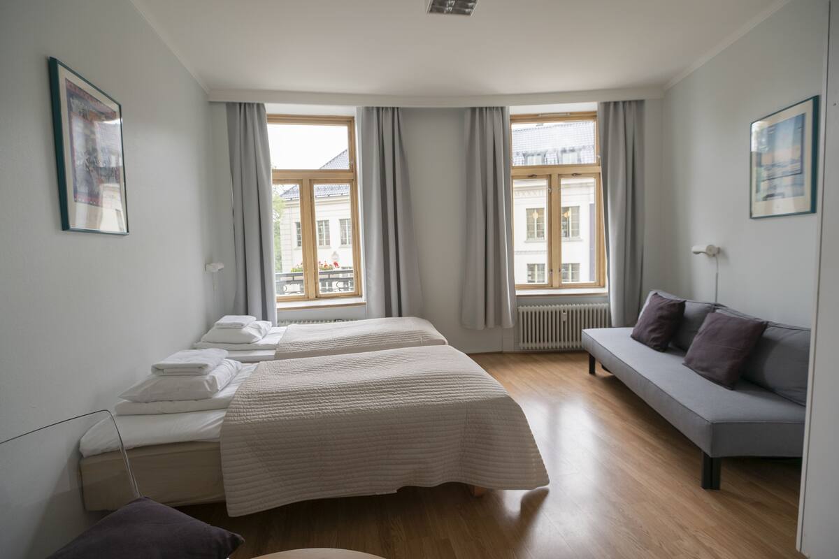 A modern hotel room with two beds, a sofa, and large windows.