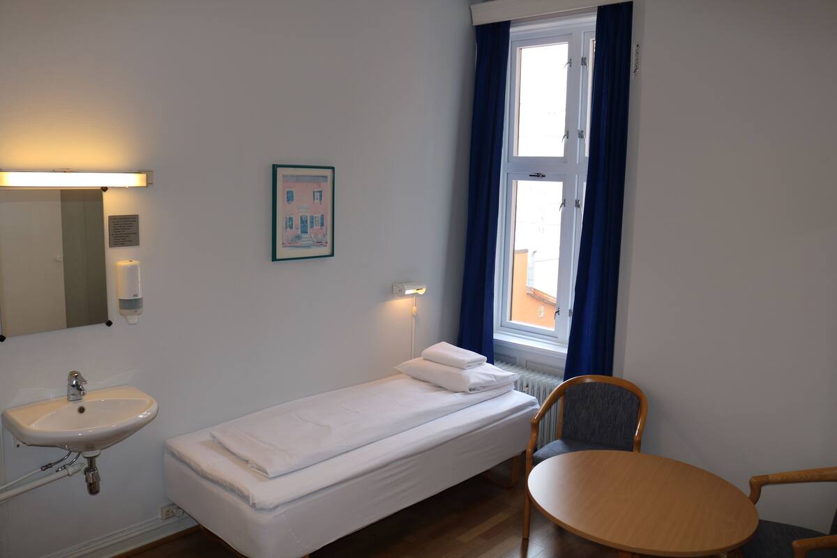 A small, clean room with a single bed, a bedside table, a chair, a window with curtains, a wall-mounted sink, and a framed picture.