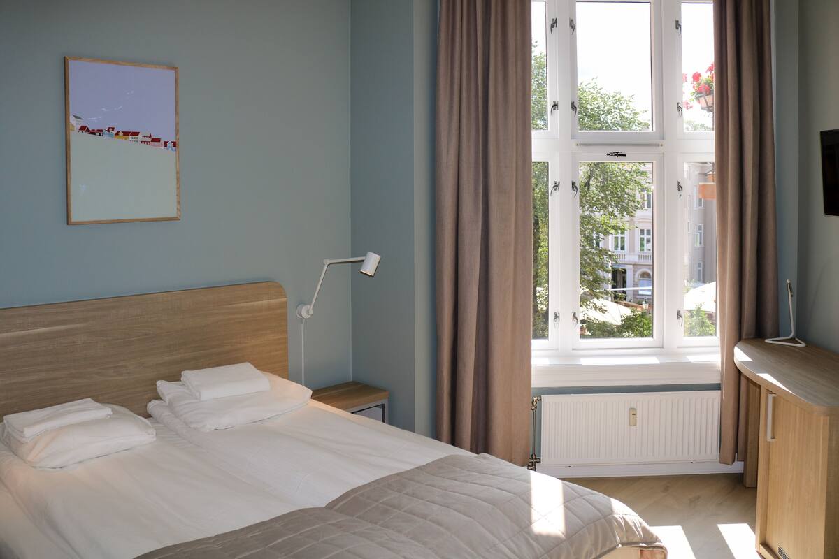 A hotel room with a bed, a bedside table, a lamp, a window with curtains, and a view of buildings outside.