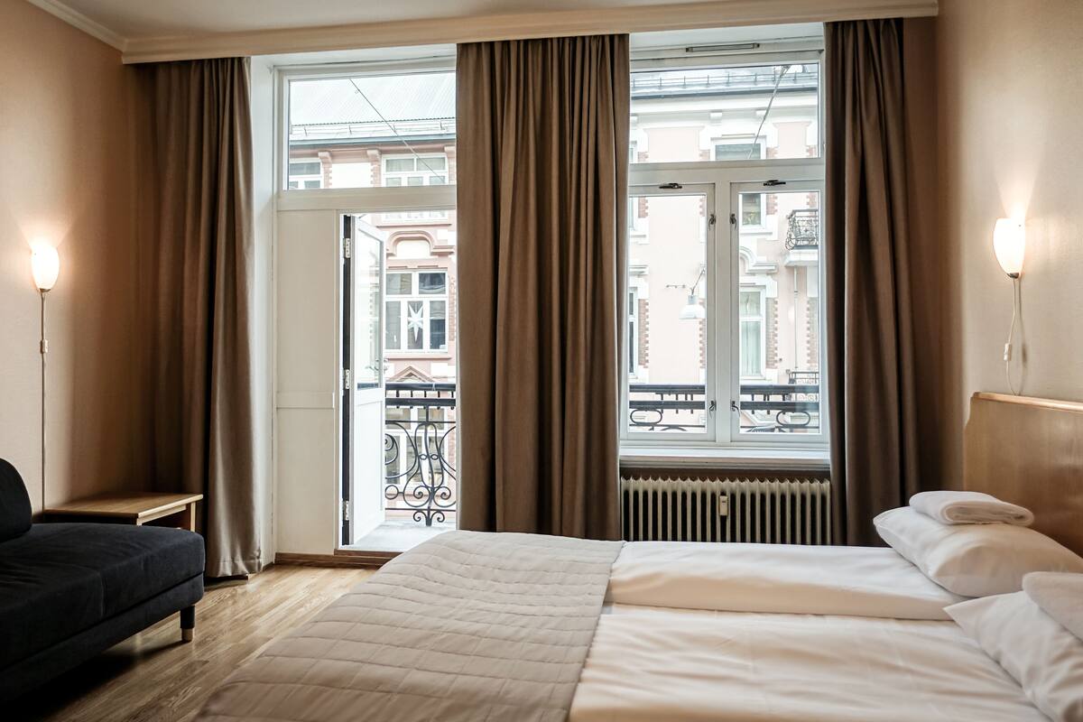 A hotel room with a bed, a chair, a window with curtains, a radiator, and a view of buildings outside.