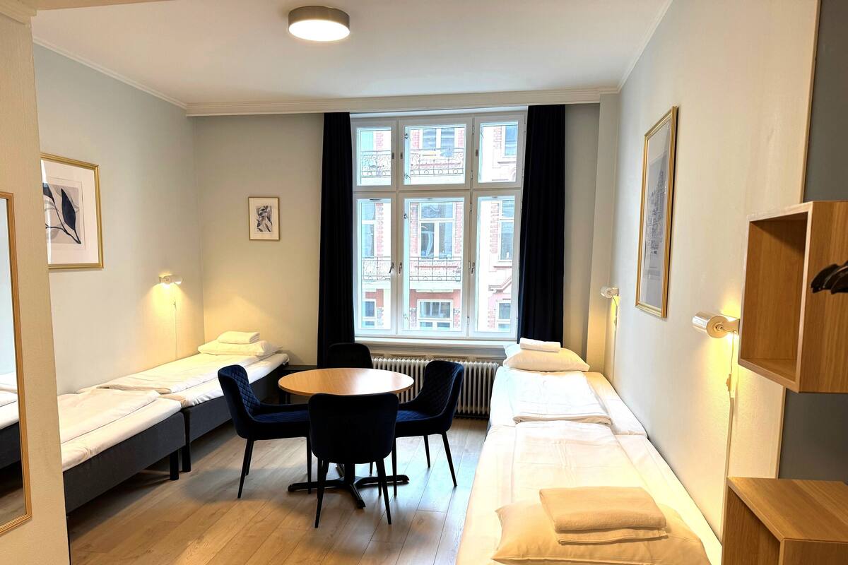 A compact hotel room with a bed, a small round table, two chairs, a radiator, and a window with a view of buildings.
