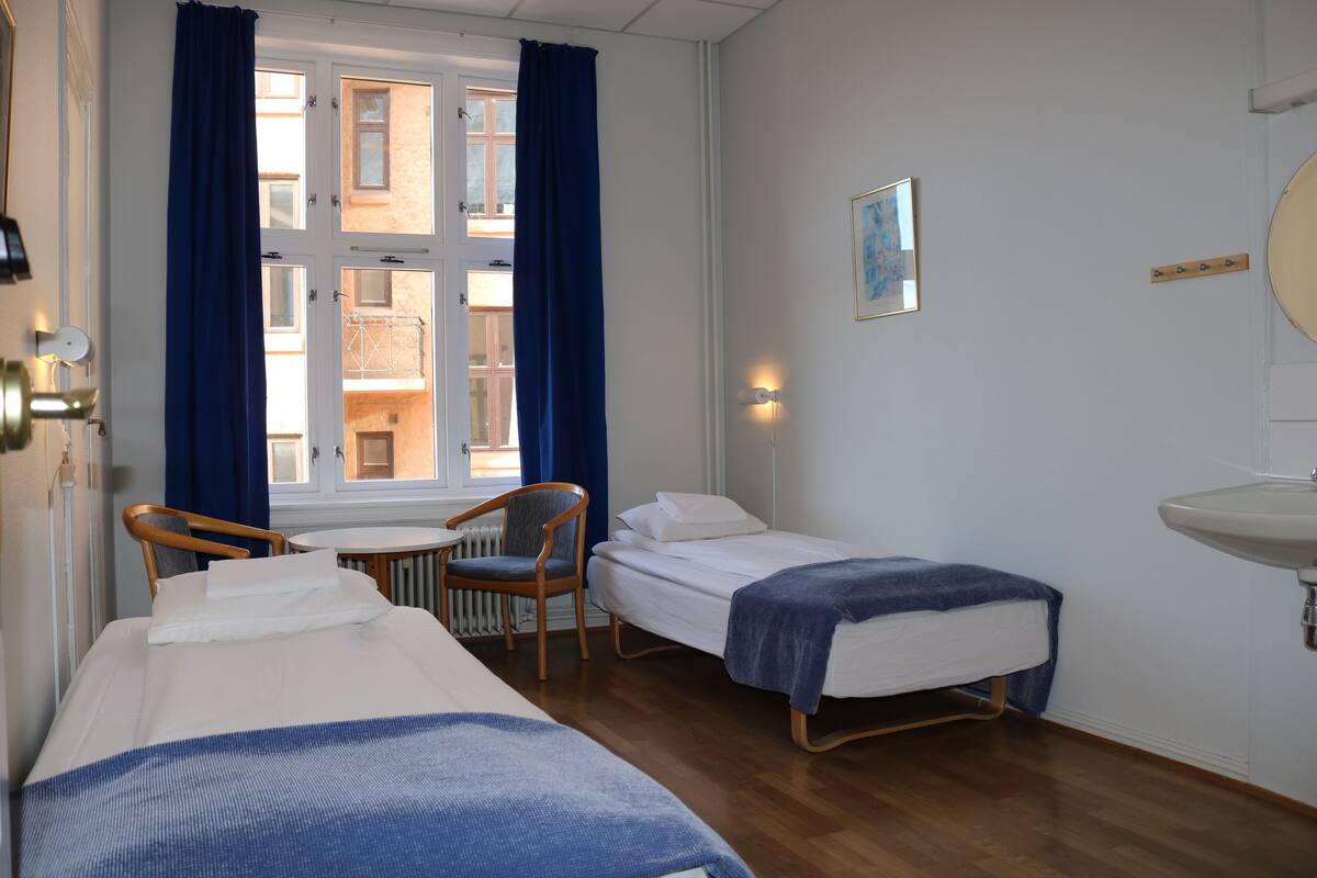 A hotel room with two single beds, a chair, a small table, a window with curtains, a wall-mounted light, and a sink.