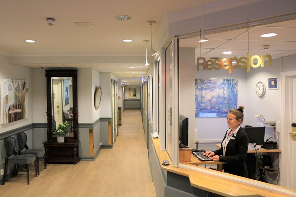 A reception area with a receptionist at the desk, a reception sign, framed artwork on the walls, and a hallway leading to other rooms. - Cochs Pensjonat (Oslo)