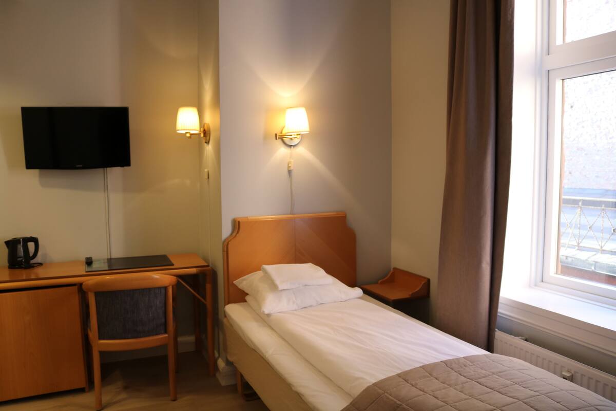 A hotel room with a bed, a desk, a chair, a TV, and two wall-mounted lamps.