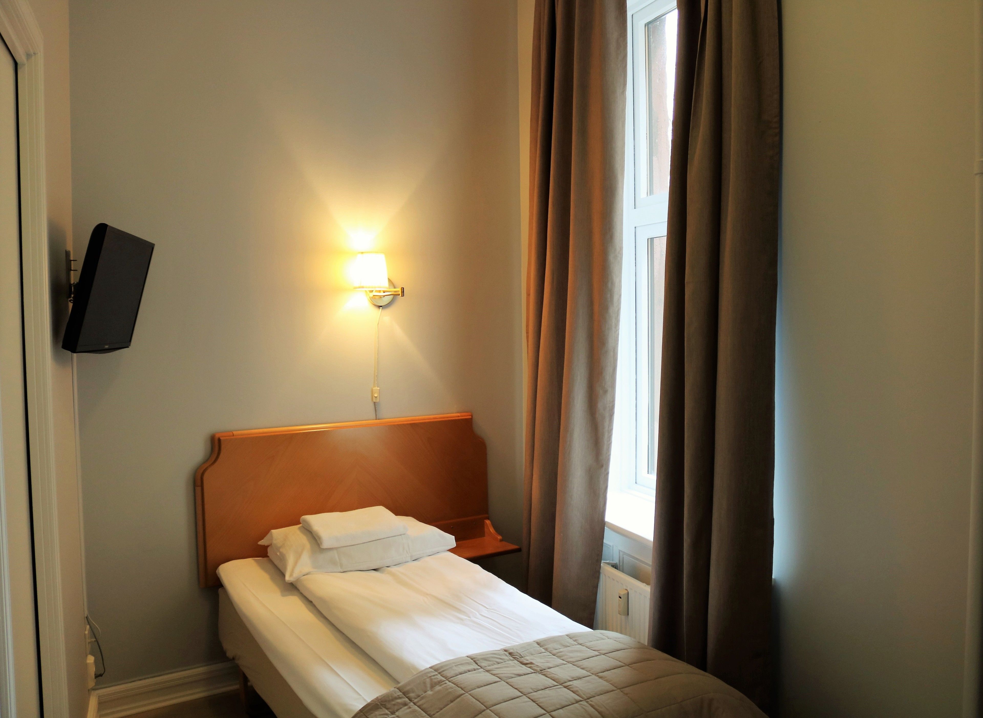 A hotel room with a bed, a wall-mounted TV, a bedside lamp, and a window with curtains.