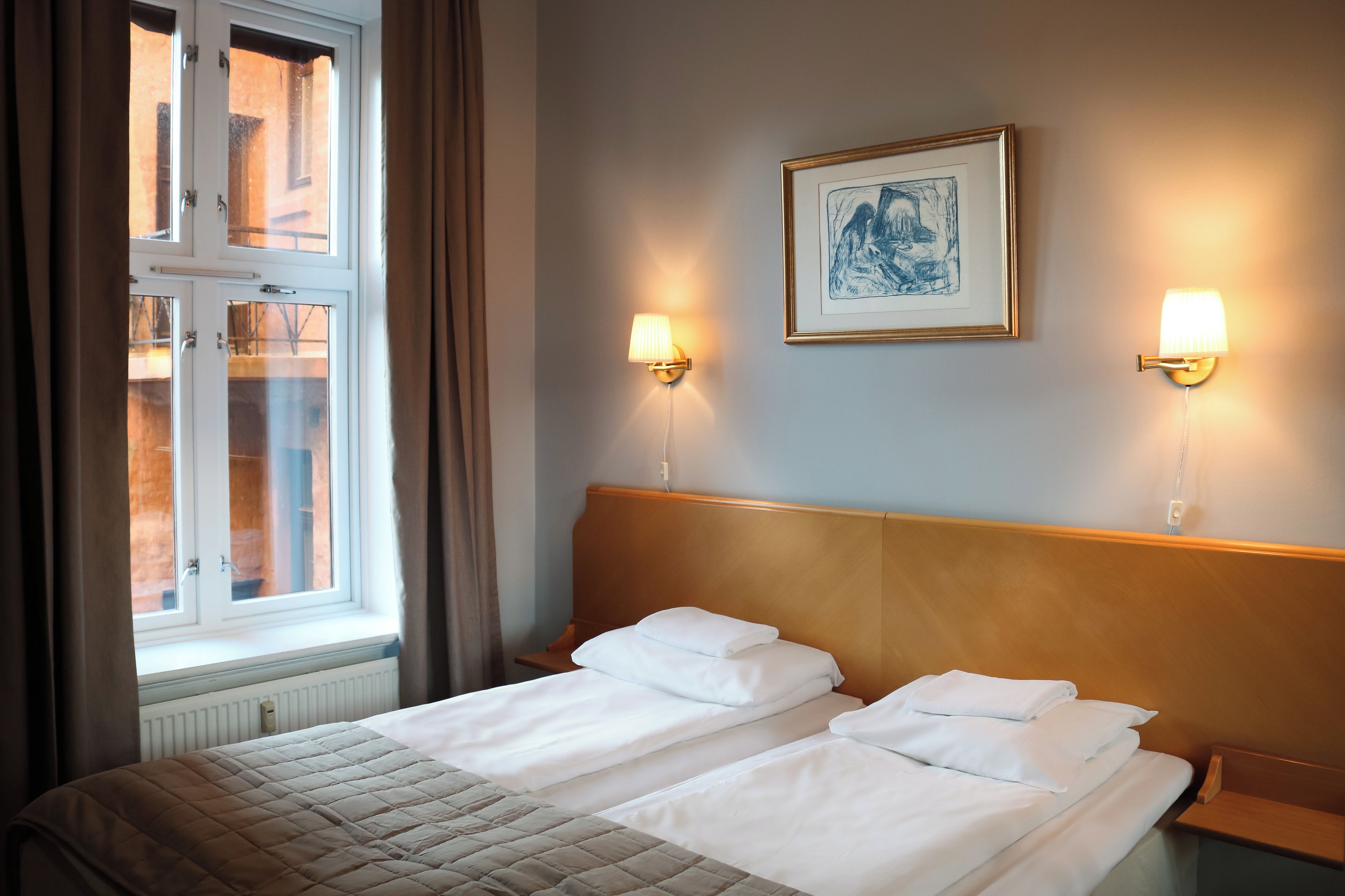 A hotel room with a double bed, two bedside lamps, a framed artwork on the wall, and a window with curtains.