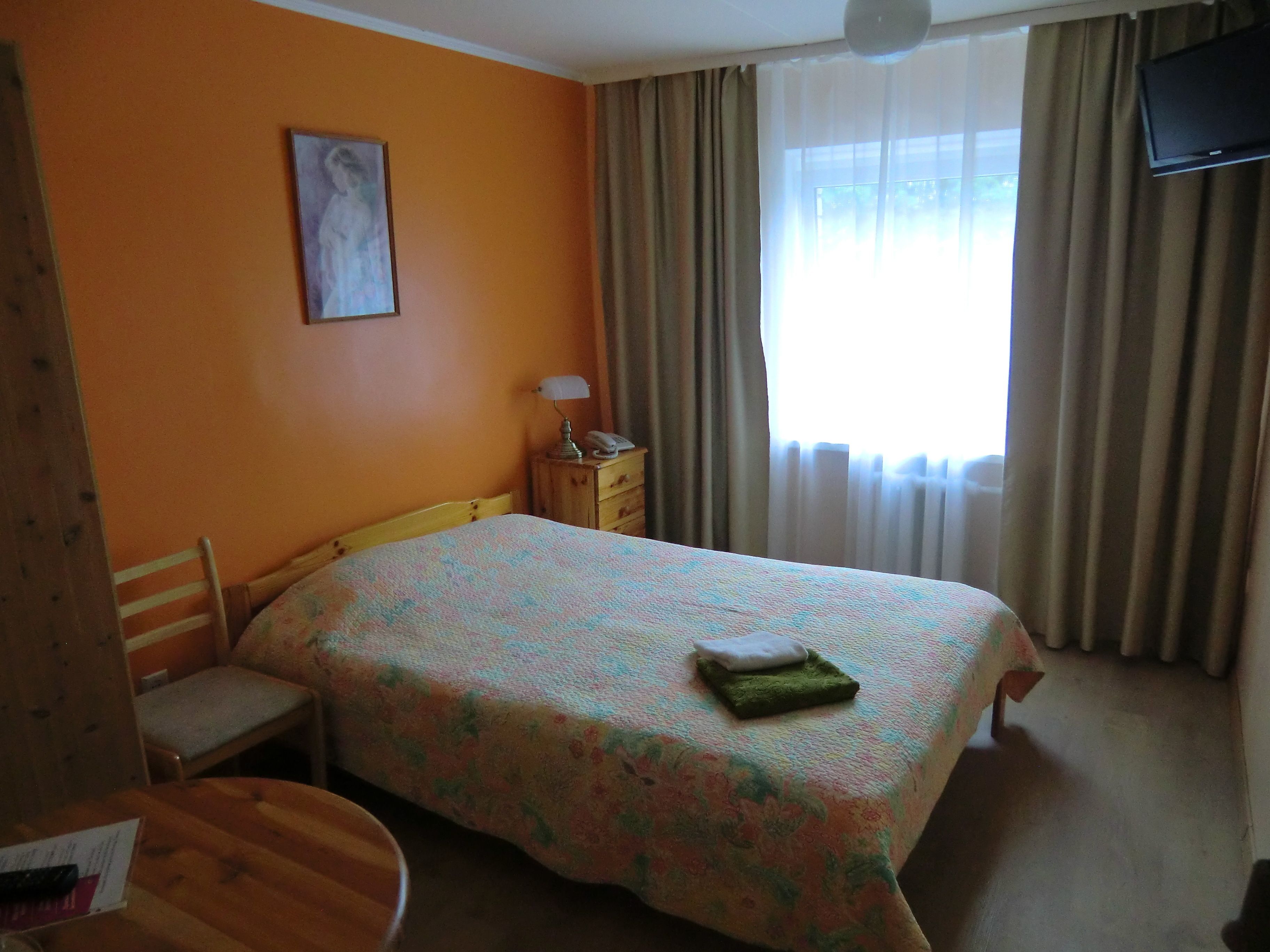 Ein Schlafzimmer mit Bett, einem Sessel, einem kleinen Tisch, einem Nachttisch, einer Lampe, einem Bild an der Wand und einem Fenster mit Vorhängen.