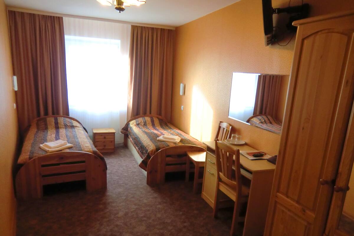 Ein Hotelzimmer mit zwei Einzelbetten, einem Schreibtisch, einem Stuhl, einem Kleiderschrank und einem Fenster mit Vorhängen.